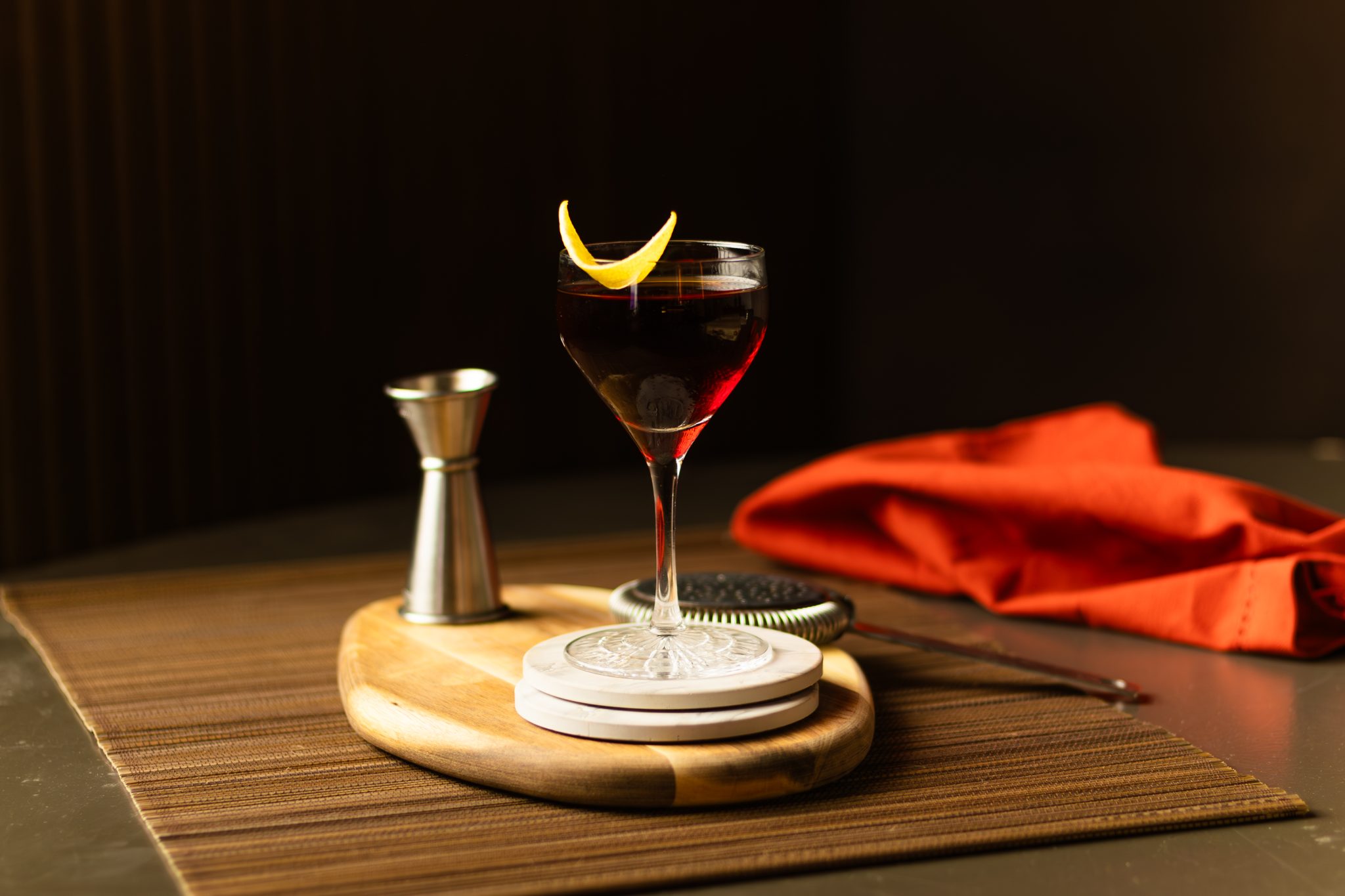 A side shot of a Remember the Maine cocktail in a coupe glass on a white coaster placed on wooden board on a brown placemat surrounded by red cloth, a strainer, and a jigger.