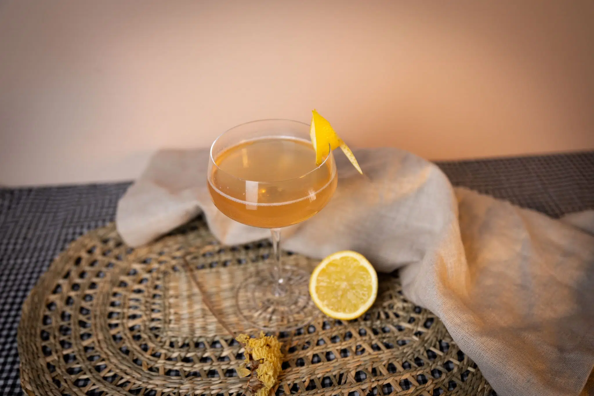 A side shot of a Romancing the Stone cocktail in a coupe glass on a straw placeemat surrounded by a beeige cloth, half a lemon, and a flower sprig