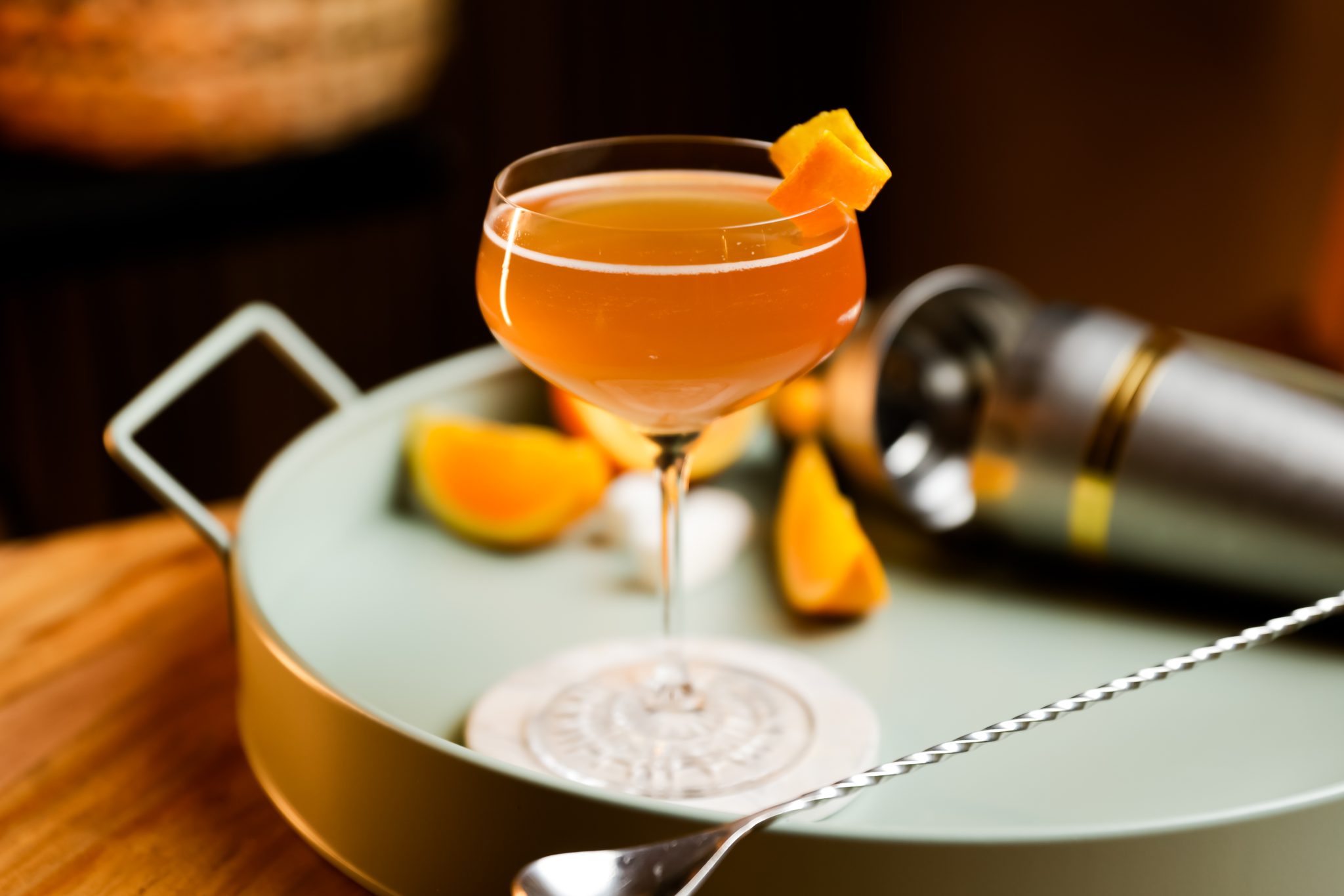 A side shot of a Roosevelt cocktail in a coupe glass on a white coaster and a turquoise tray with three orange wedge, a cocktail shaker and a bar spoon behind