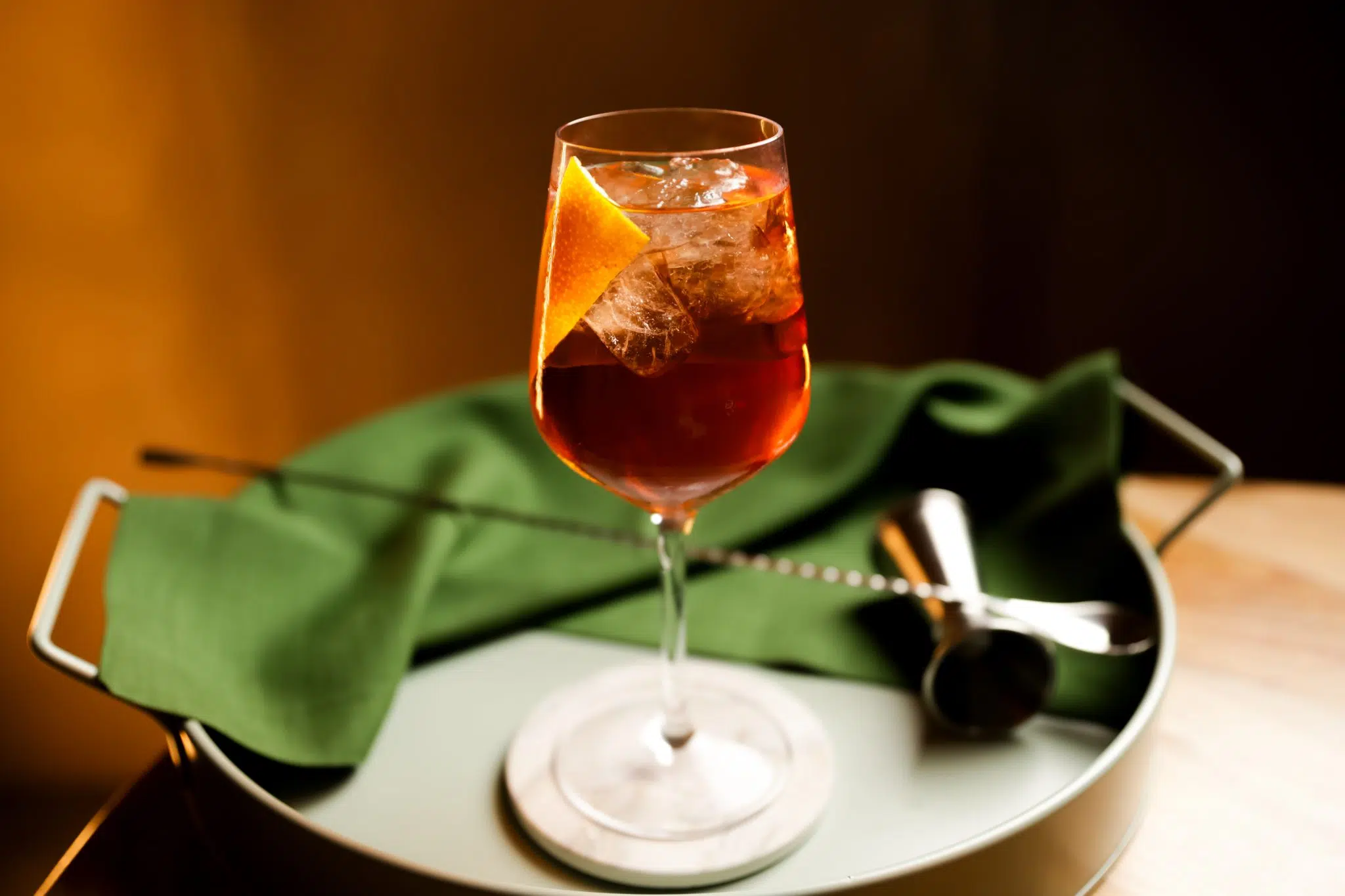 A side shot of a Rosé Aperol Spritz cocktail in a wine glass on a white coaster surrounded by a jigger, a bar spoon and a green cloth