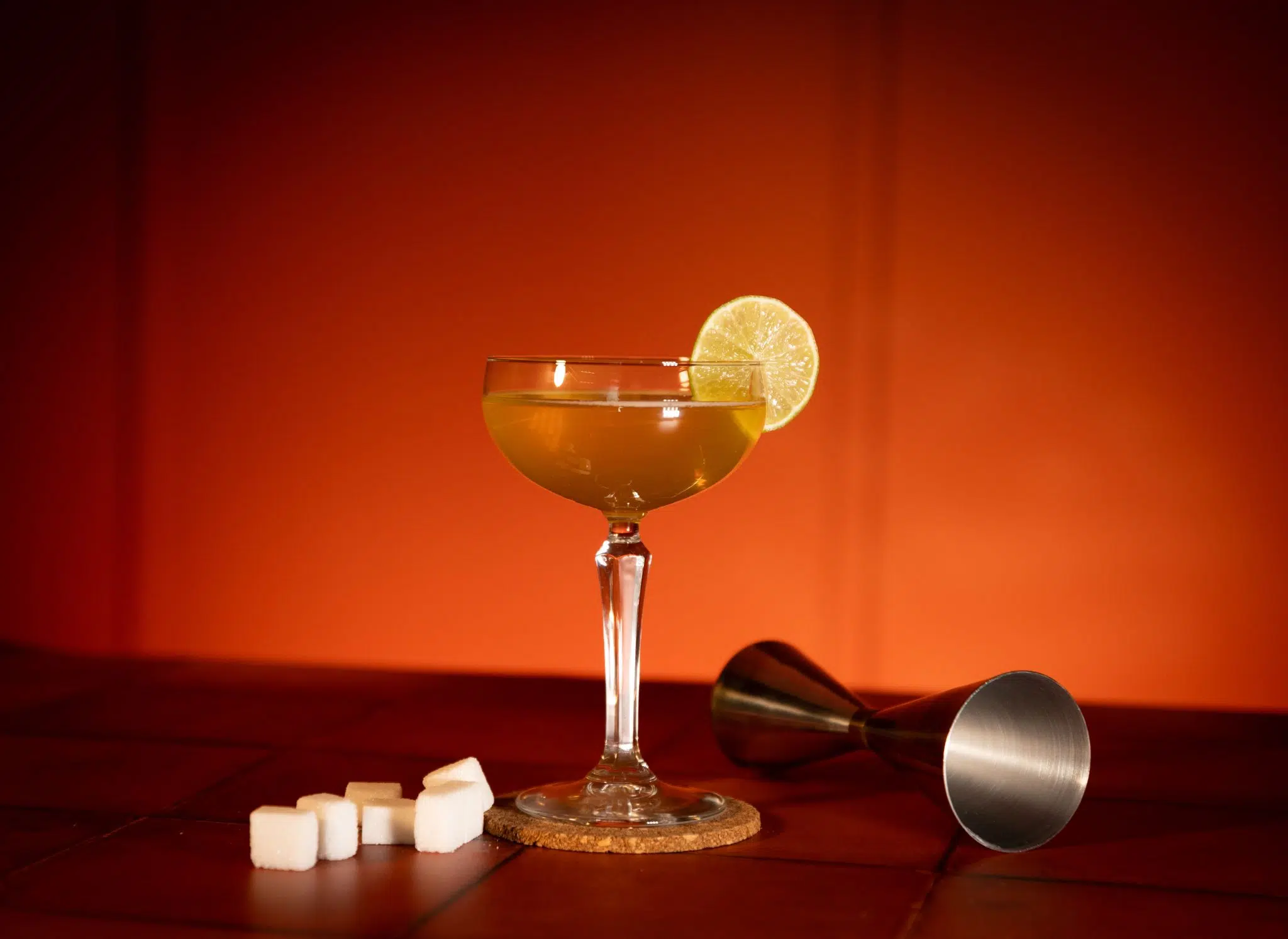 A side shot of a Royal Bermuda Yacht Club in a coupe glass on a cork coaster surrounded by a jigger and sugar cubes