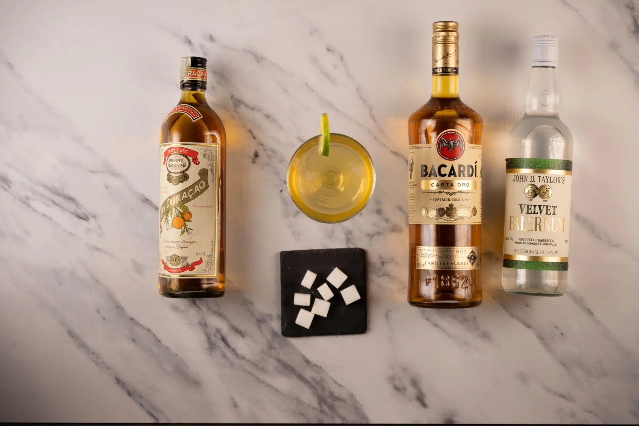 A Royal Bermuda Yacht Club cocktail, shot from above, in a coupe glass on a white marmol surface surrounded by Gold rum, Falernum liqueur, orange curaçao, lime and sugar cubes