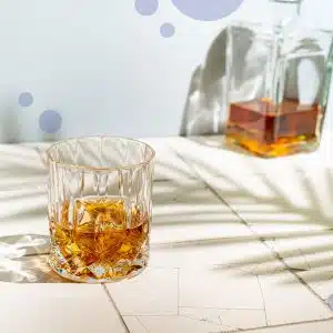 Old-fashioned glass filled with rum on a white floor with a transparent bottle with rum in front of a light blue wall.