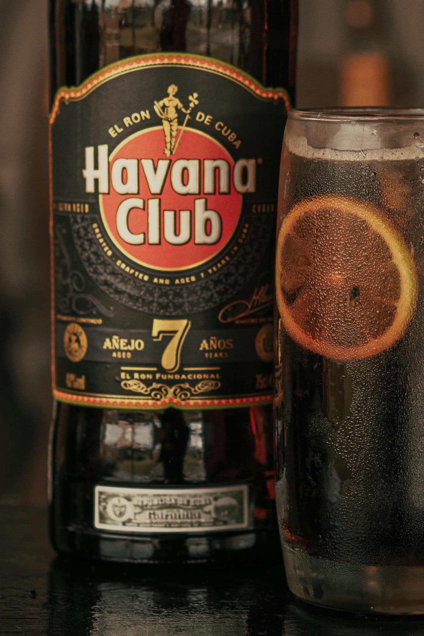 A rum bottle of Havana Club Añejo 7 Años next to a rum drink with a slice of orange inside.