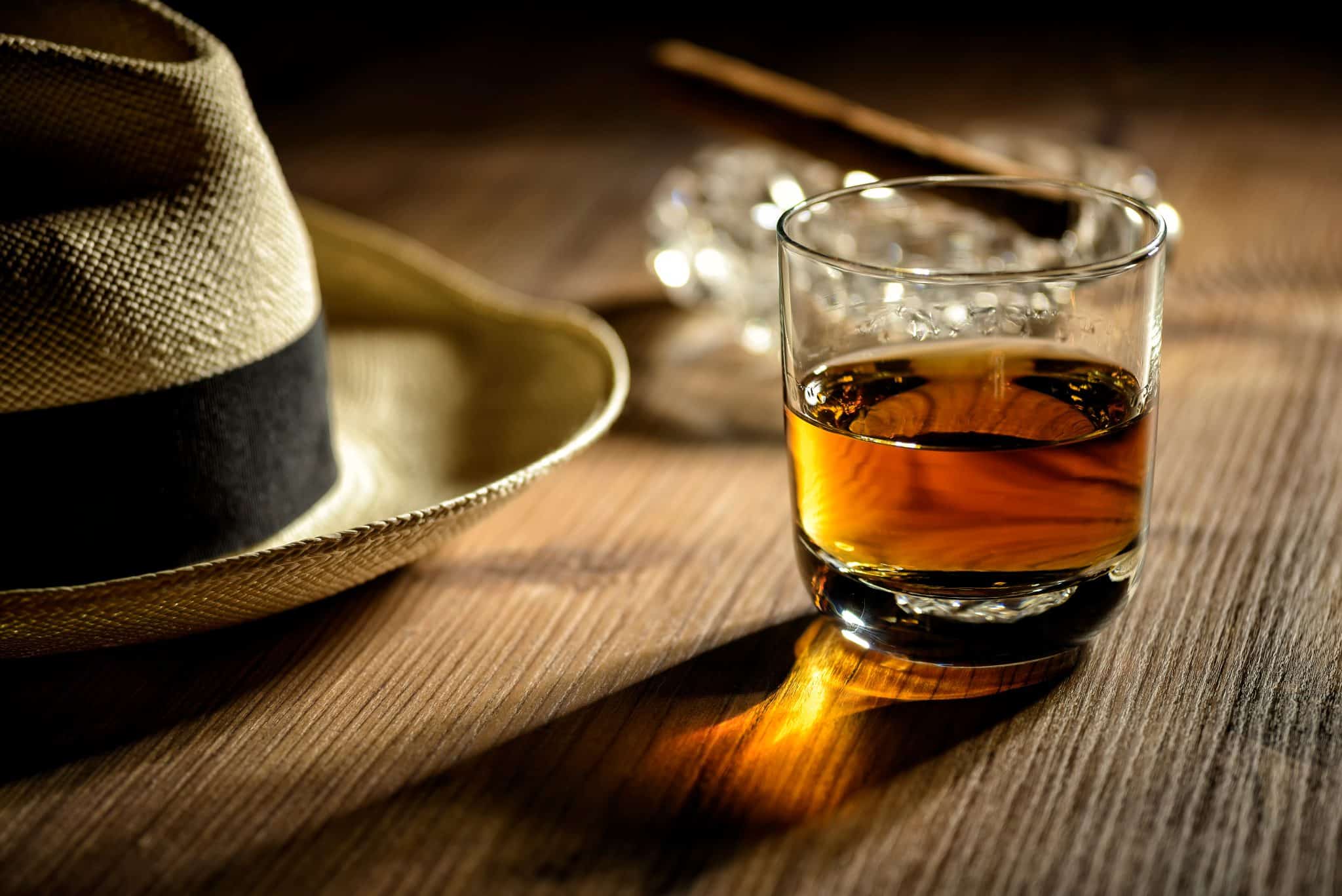 A glass of neat rum, a straw hat, and a cheroot on a wooden surface.