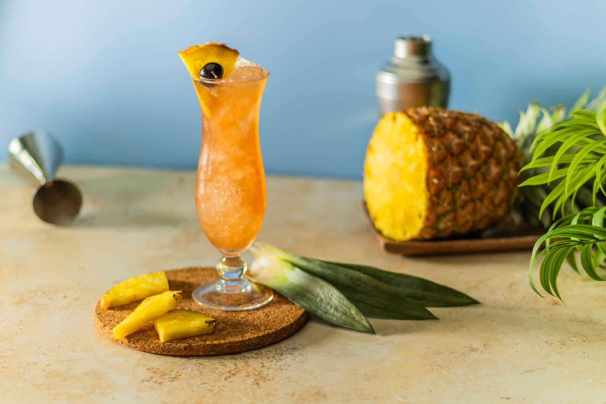 A Rum Runner cocktail, shot from a side, in a highball glass with three pineapple wedges on a wooden plate surrounded by a jigger, a shaker and a pineapple fruit