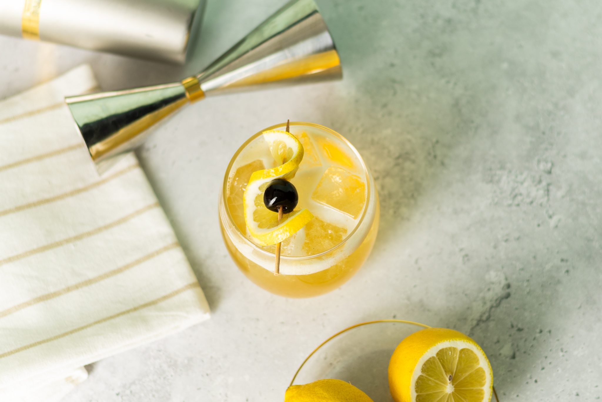 A Rum Sour cocktail, shot from above, in a rocks glass on a white table with a jigger, a white cloth, and a plate with two lemons around.