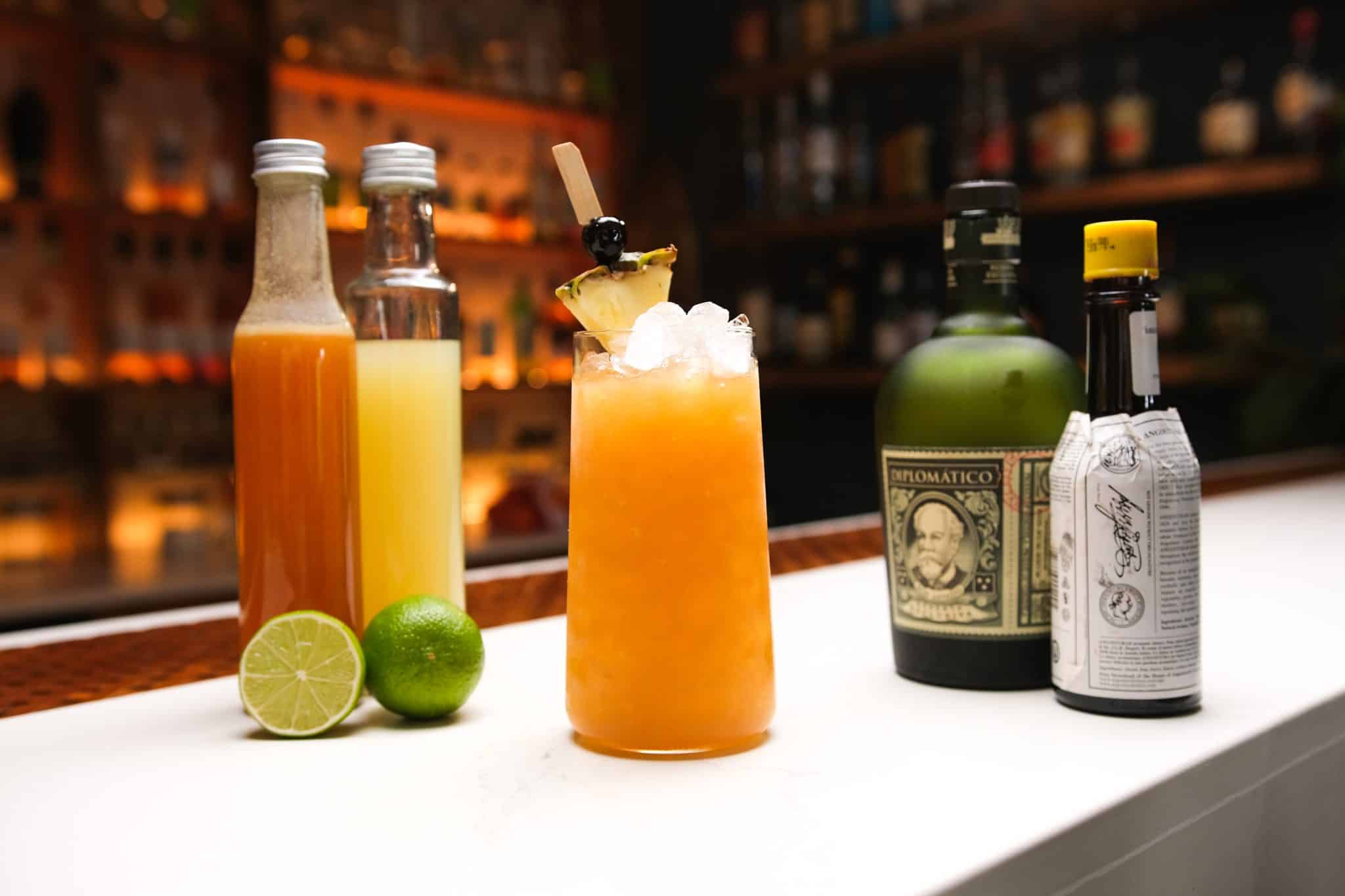 Pineapple juice, orange juice, Angostura Bitters, Dark Rum and two limes laid out on a white bar table.