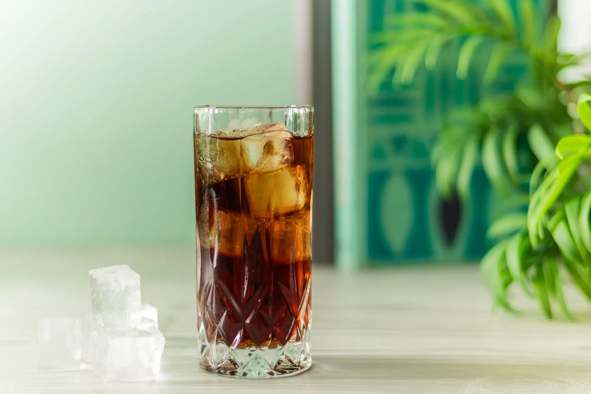 A side shot of a Rum and Coke cocktail in a highball glass with 3 ice cubes on the side and turquoise background color
