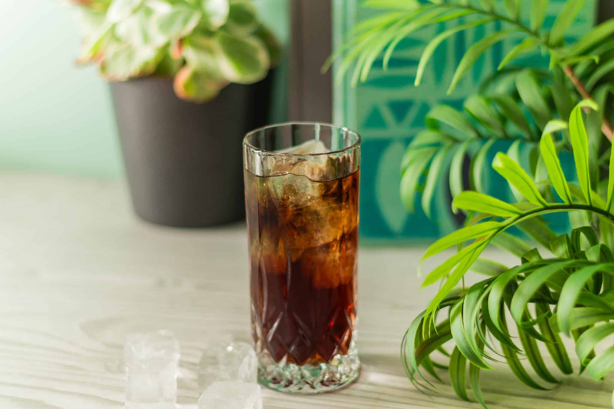 A side shot of a Rum and Coke cocktail in a highball glass on a table with a plant behind and turquoise background color