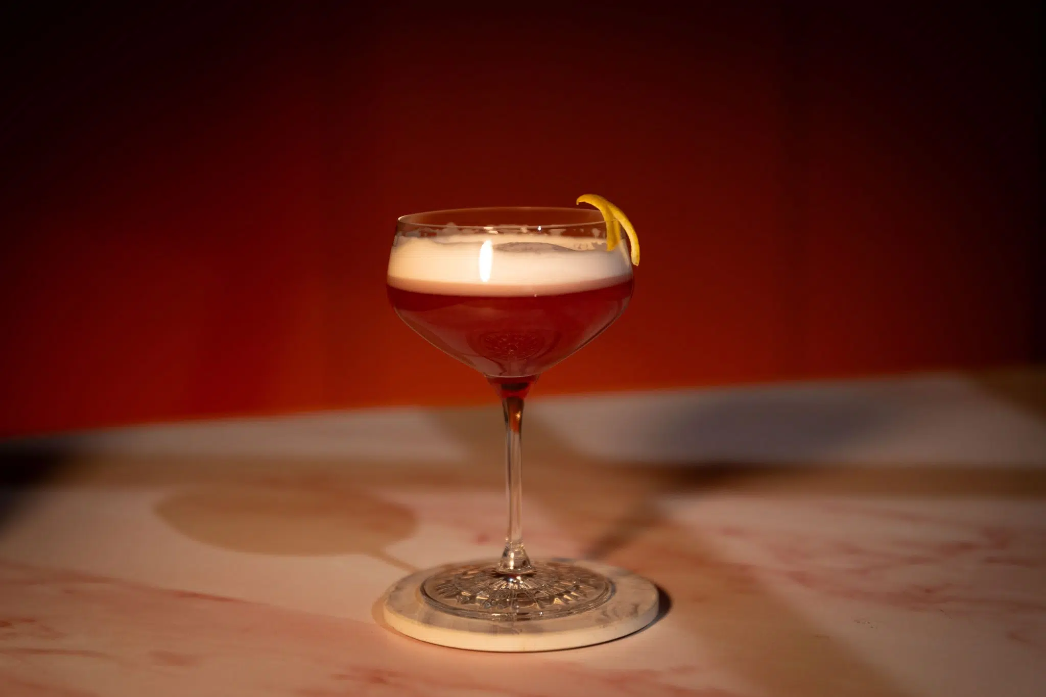 A side shot of a Rumbera cocktail in a coupe glass on a wooden coaster placed on a wooden surface in front of a red background