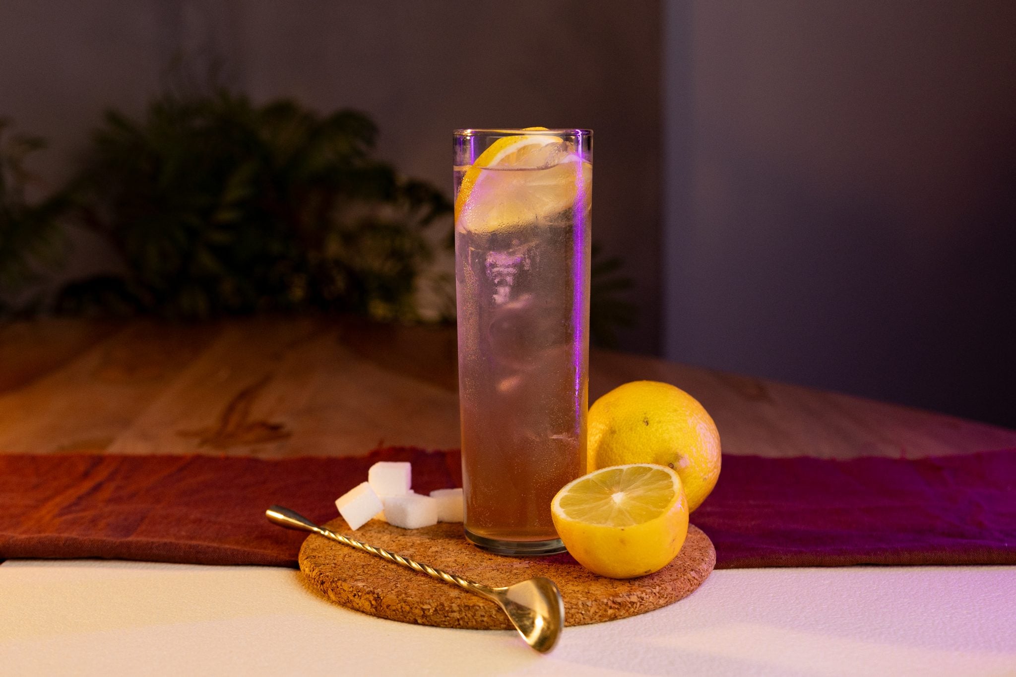 A side shot of a Rye Collins cocktail in a highball glass on a cork tray surrounded by two lemons, a bar spoon and four sugar cubes