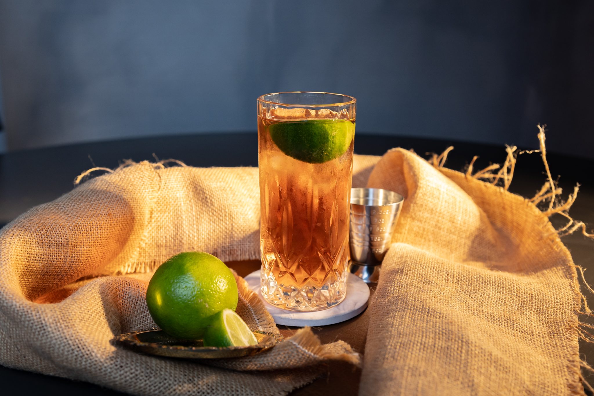 A side shot of a Rye and Ginger cocktail in a highball glass on a white coaster surrounded by beige cloth, a lime, a lime wedge and a jigger