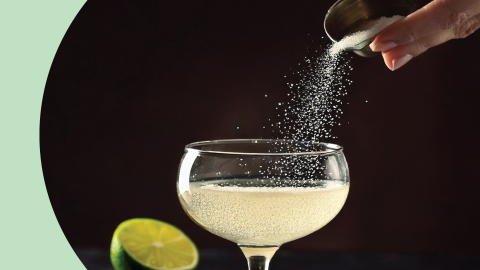 A shot of a hand pouring salt into a cocktail in a coupe glass in front of a black background