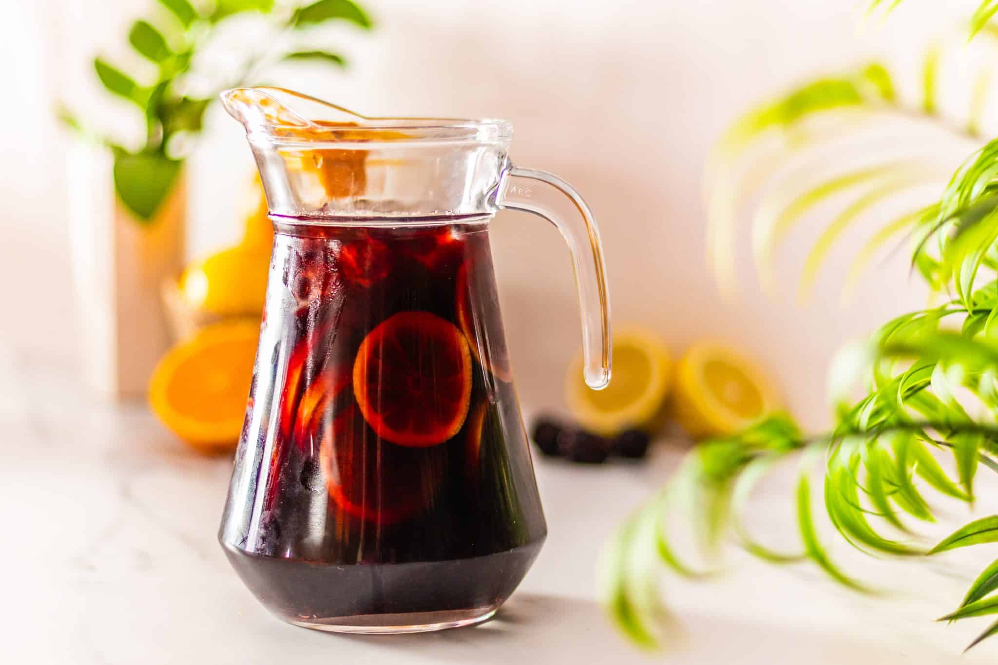 A Sangria Drink in a pitcher on a white table with some oranges behind and some plant leaves on the side.
