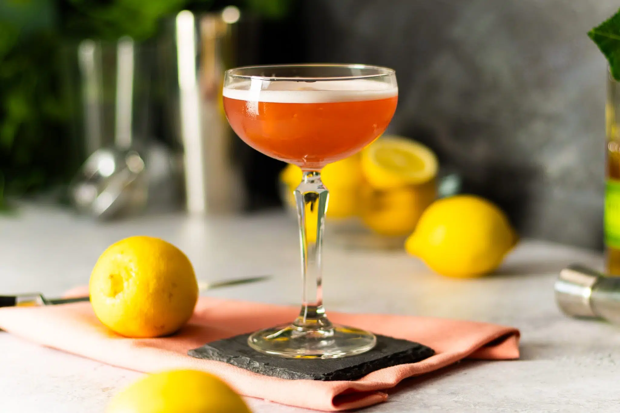 A side shot of a Scofflaw cocktail in a cocktail glass on a black stone coaster placed on a salmon cloth on a white table surrounded by five lemons, a knife, a jigger, and a shaker, in front of a grey wall.