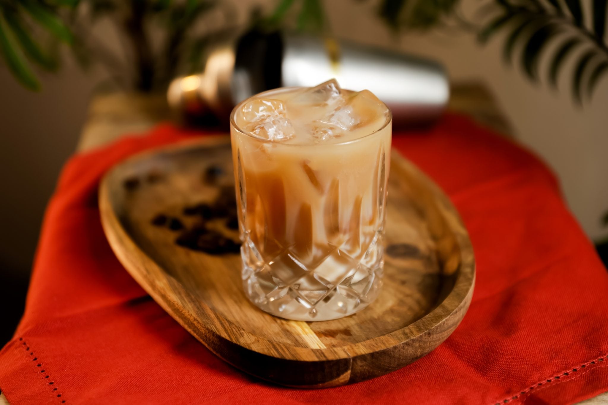A side shot of a Shaft cocktail in an Old Fashioned glass on a wooden tray with coffee beans and a cocktail shaker behind, placed on a red cloth