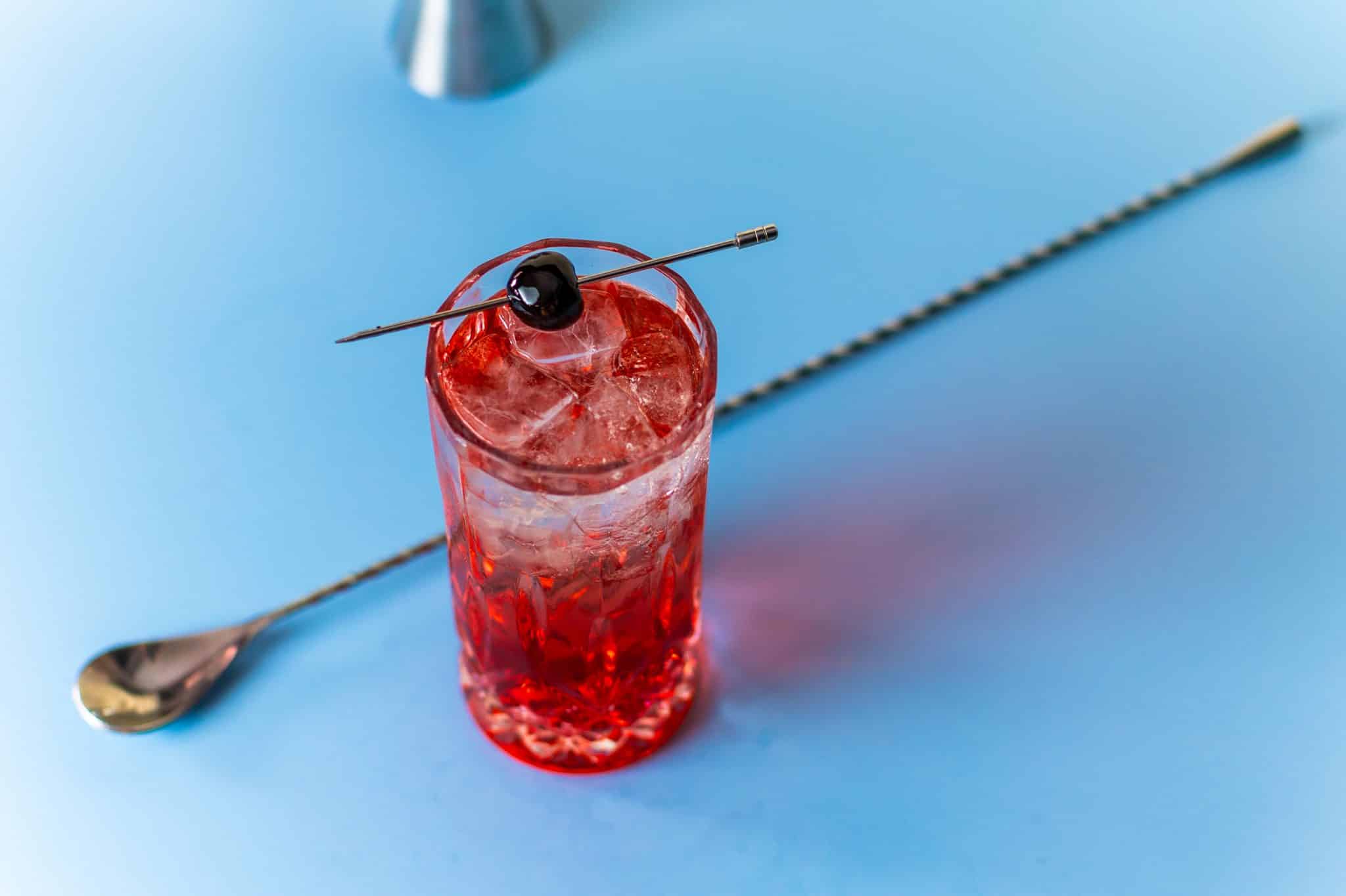 A Shirley Temple mocktail, shot from above, in a highball glass with a bar spoon on a side, placed on a blue table.