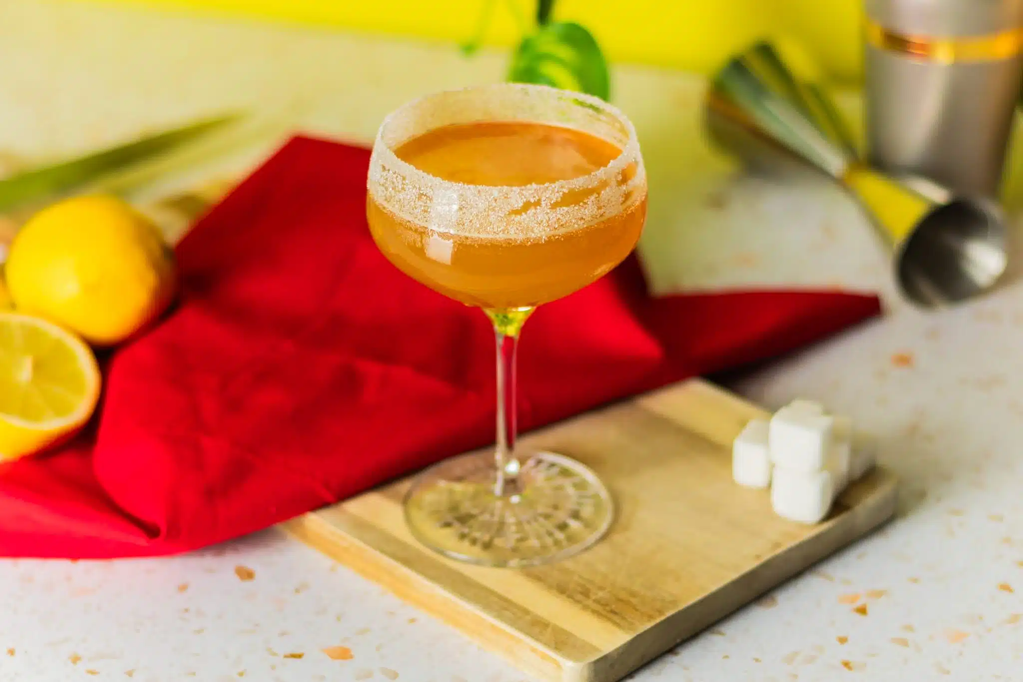A side shot of a Sidecar cocktail in a cocktail glass on a wooden board placed on a white marmol table with a red cloth, four sugar cubes, two lemons, a jigger, and a shaker around.