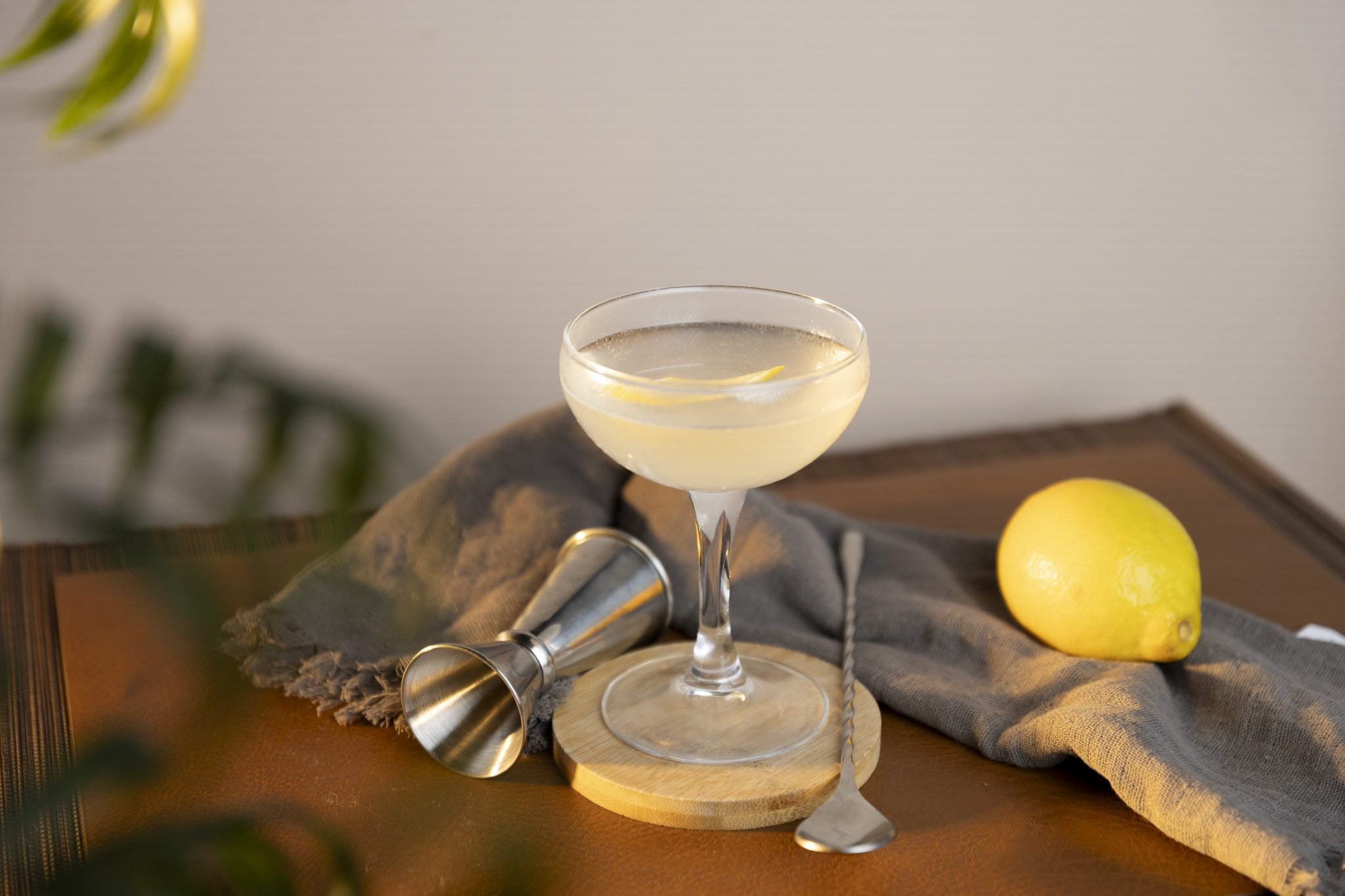 A side shot of a Silver Bullet drink in a coupe glass on a wooden coaster placed on a wooden surface surrounded by a jigger, a bar spoon, a lemon and a grey cloth