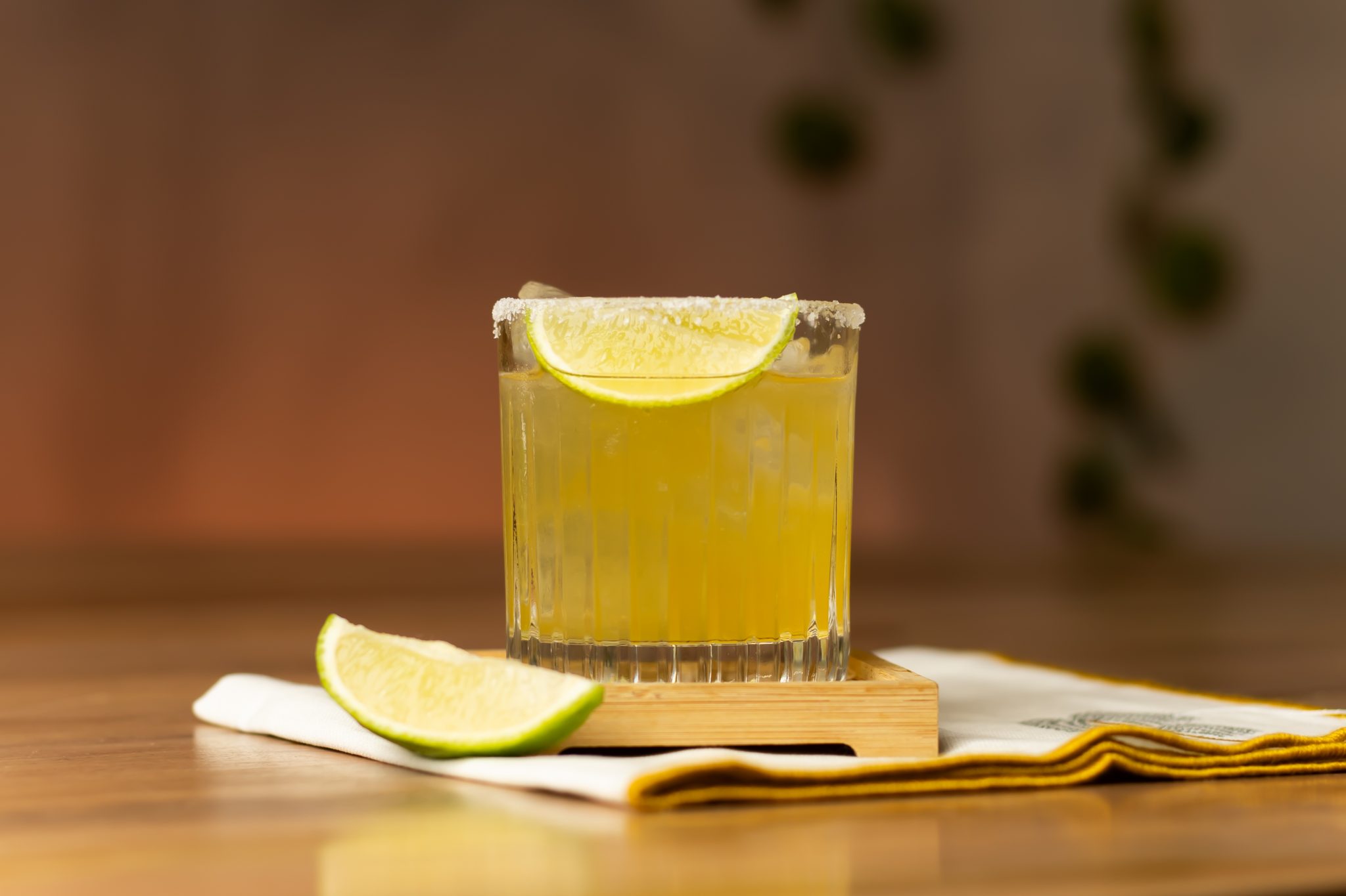 A side shot of a Skinny Margarita cocktail in an old fashioned glass on a coaster placed on a wooden table with a white cloth under the coaster and a lime wedge on one side