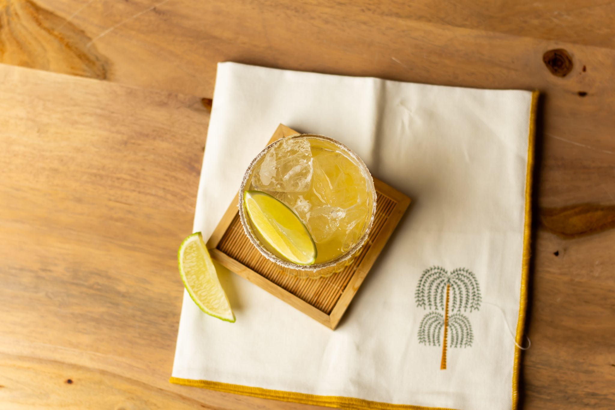 A Skinny Margarita cocktail, shot from above, in an old fashioned glass on a coaster placed on a wooden table with a white cloth under the coaster and a lime wedge on one side.