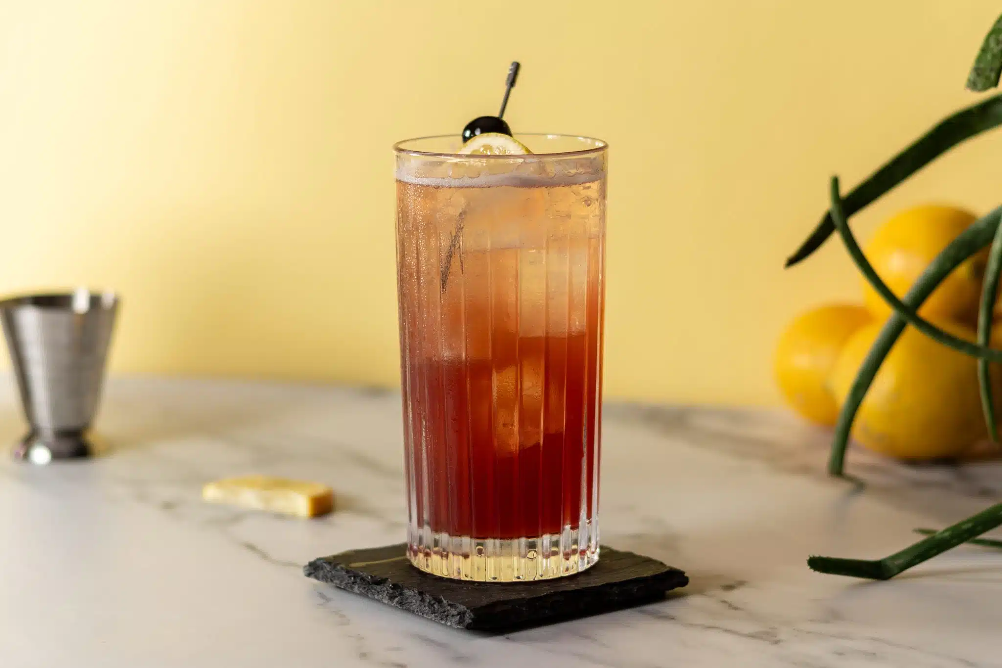 A side shot of a Sloe Gin Fizz cocktail on a black stone plate placed on a white marmol surface with a jigger and oranges on the background