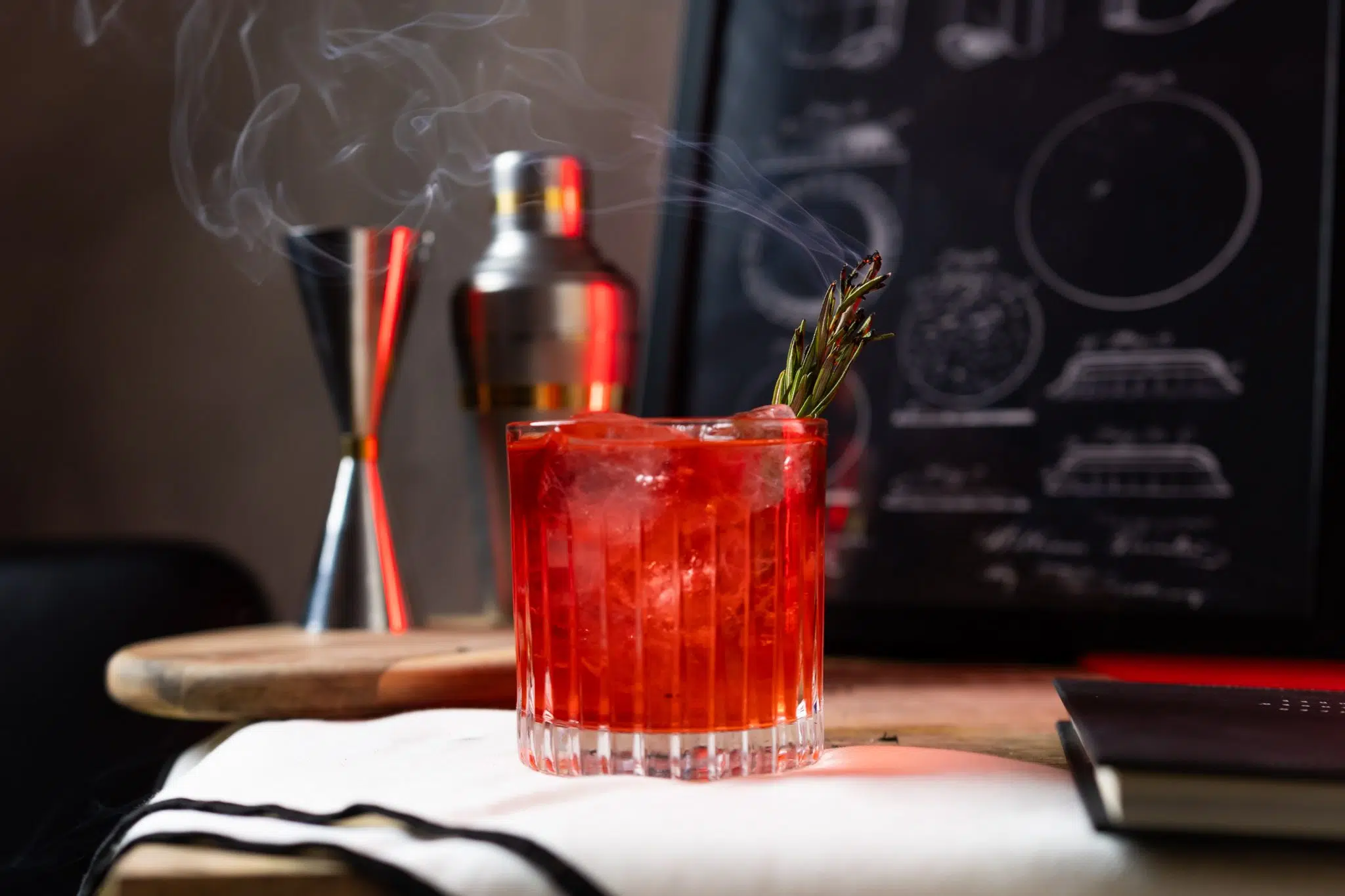 A side shot of a Smoke on the Water cocktail in an old fashioned glass on a white cloth in front of a jigger, a cocktail shaker, and a black frame