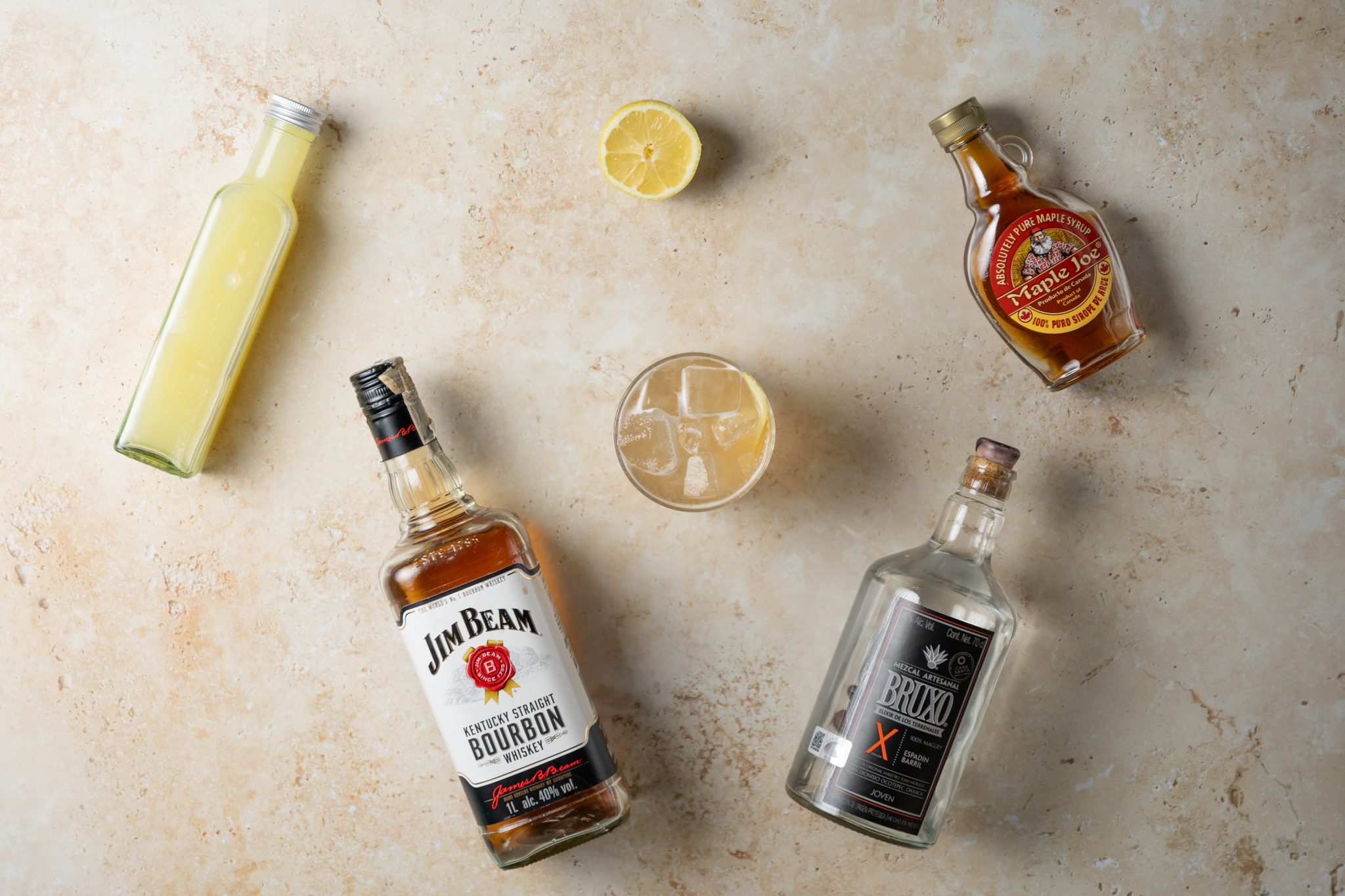 A Smokey Robinson cocktail, shot from above, in an old fashioned glass on a beige surface surrounded by Whiskey, Mezcal, lemon juice, maple syrup, and lemon