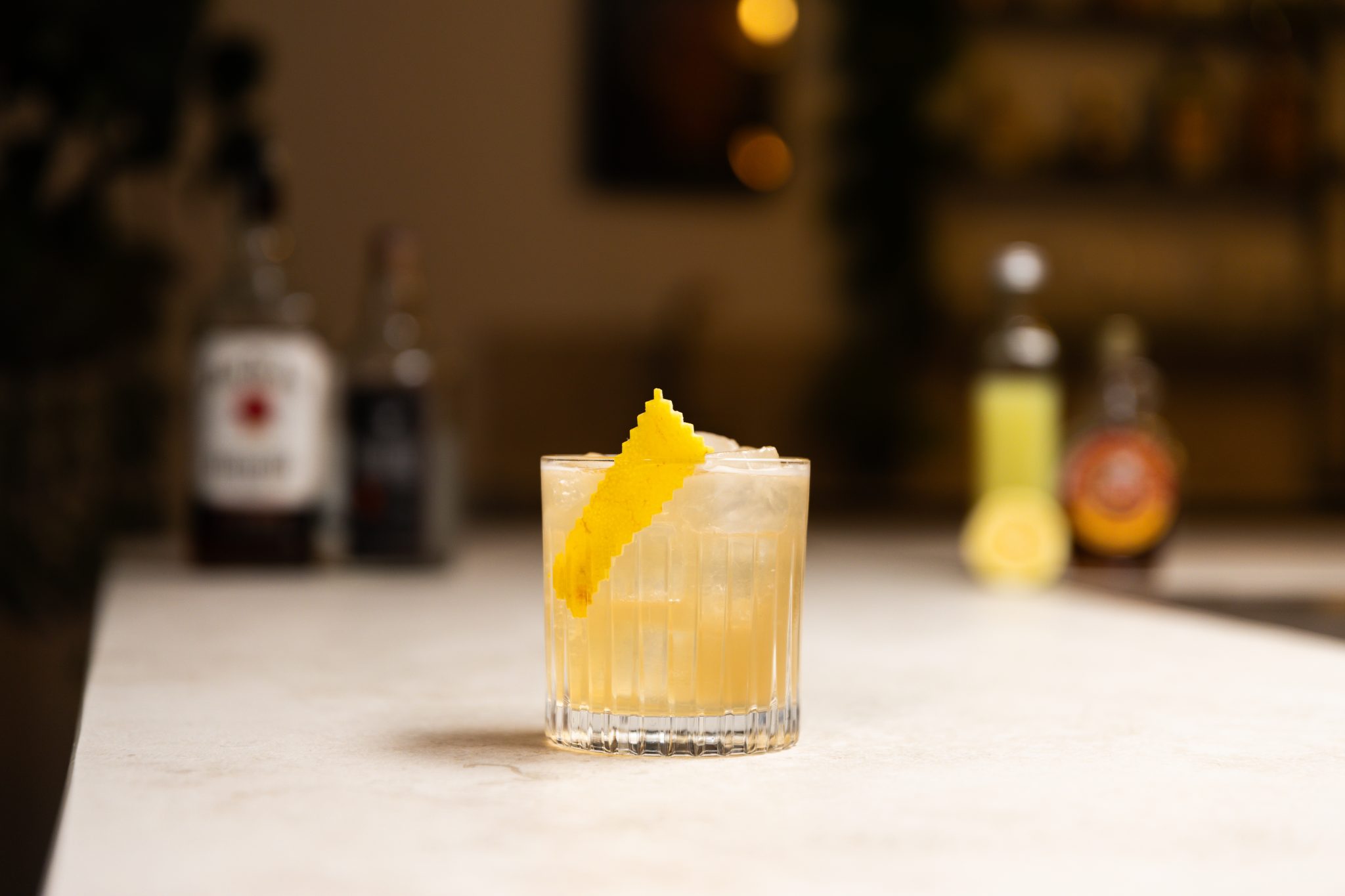 Whiskey, Mezcal, lemon juice, maple syrup, and lemon laid out on a white bar table