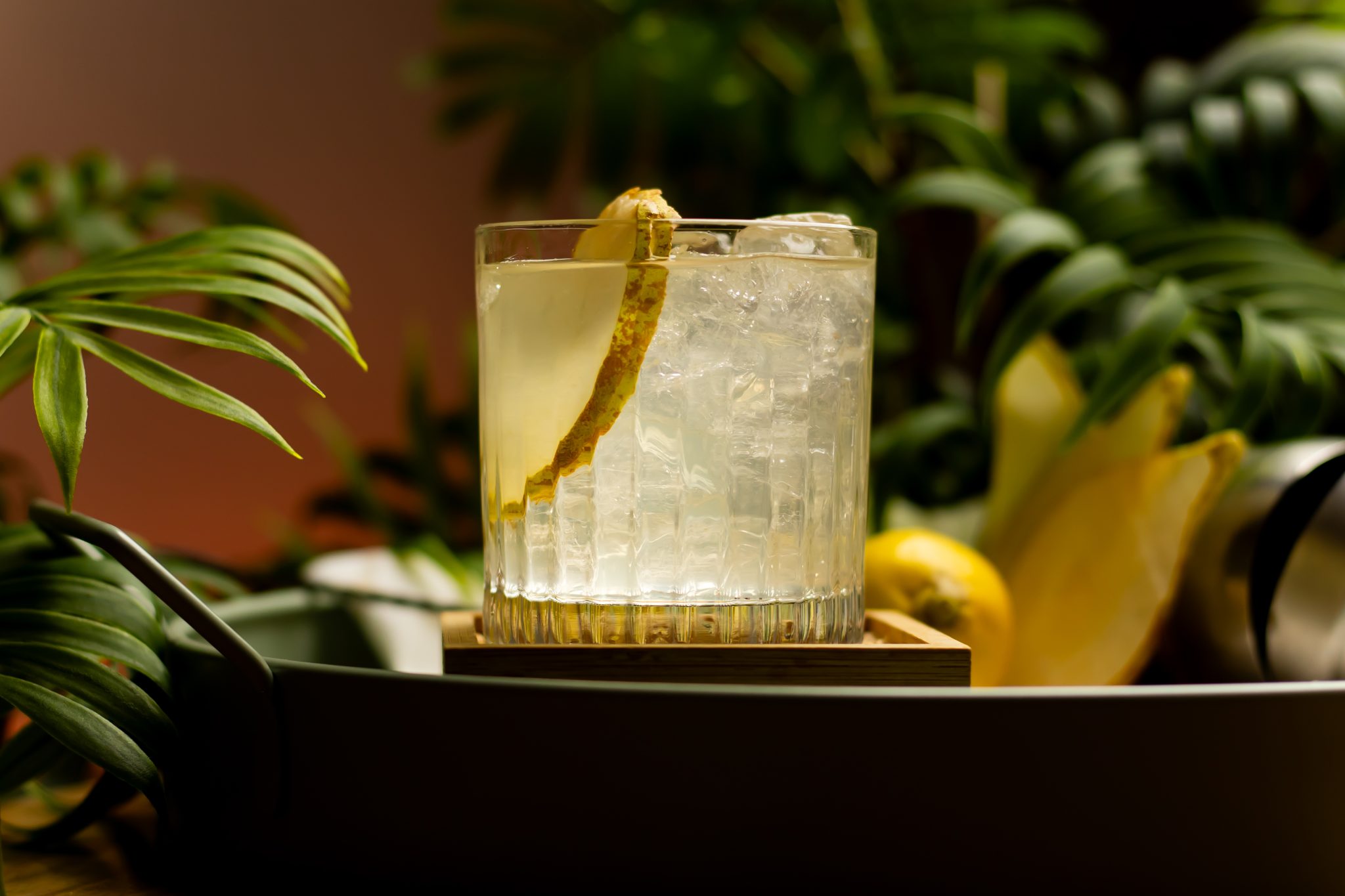 A side shot of a Smoky Pear cocktail in an old fashioned glass on a wooden coaster and a turquoise tray surrounded by a lemon, a shaker and plants