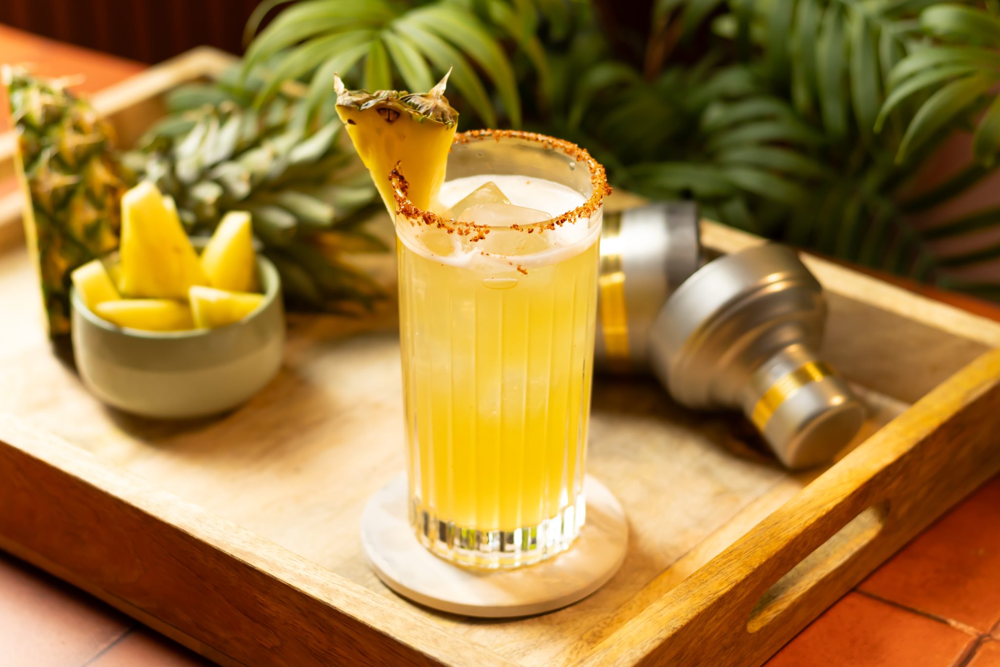 A side shot of a Smoky Pineapple juice in a highball glass on a white coaster placed on a wooden tray surrounded by a cocktail shaker, a bowl with pineapple slices, and a pineapple piece