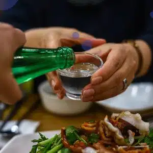 Round shot glass, in the hands of a person, being filled from a green glass bottle with soju above of a table full serve with food.