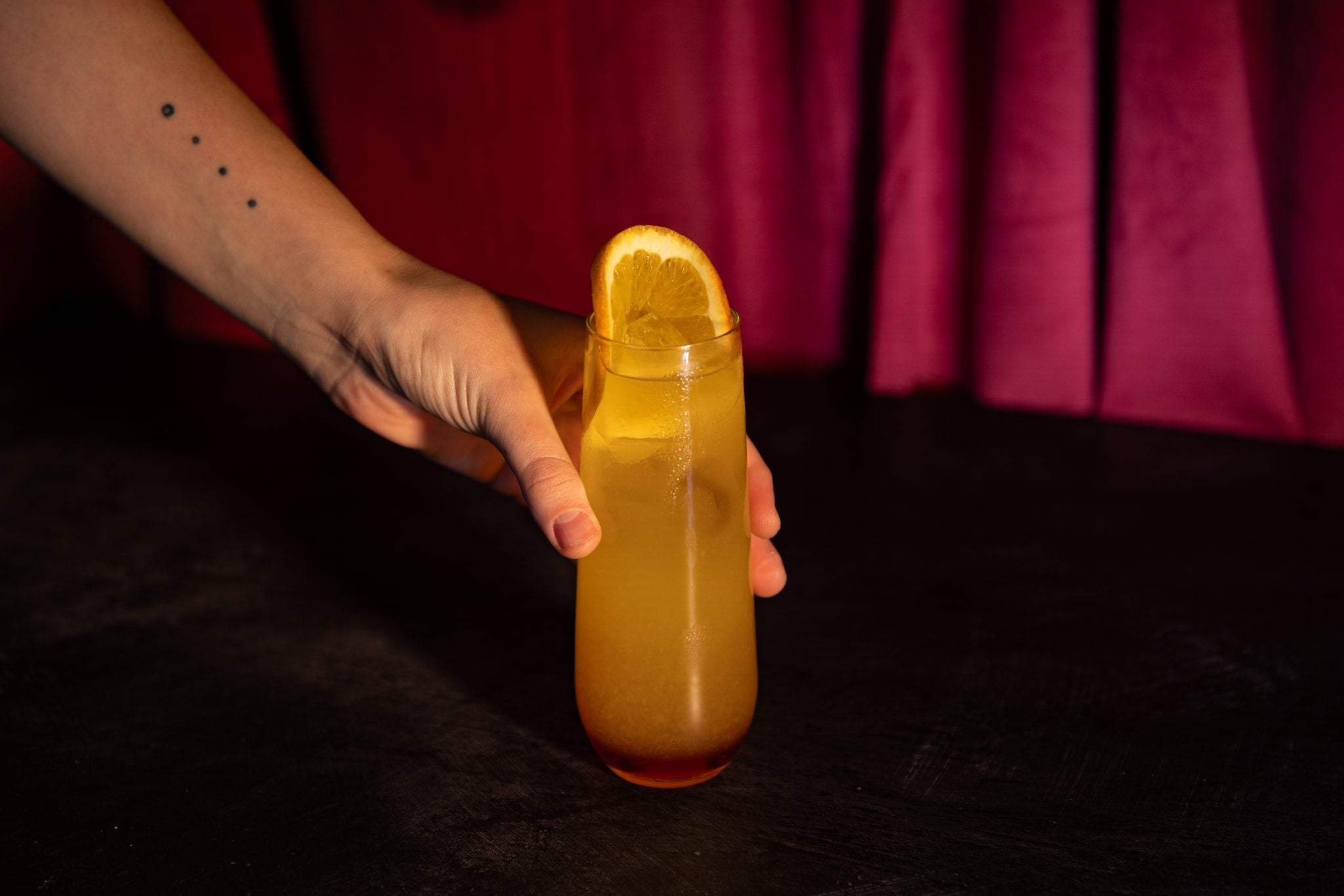 A side shot of a Soju Sunrise cocktail in a highball glass on a black surface holded by a hand in front of a red curtain