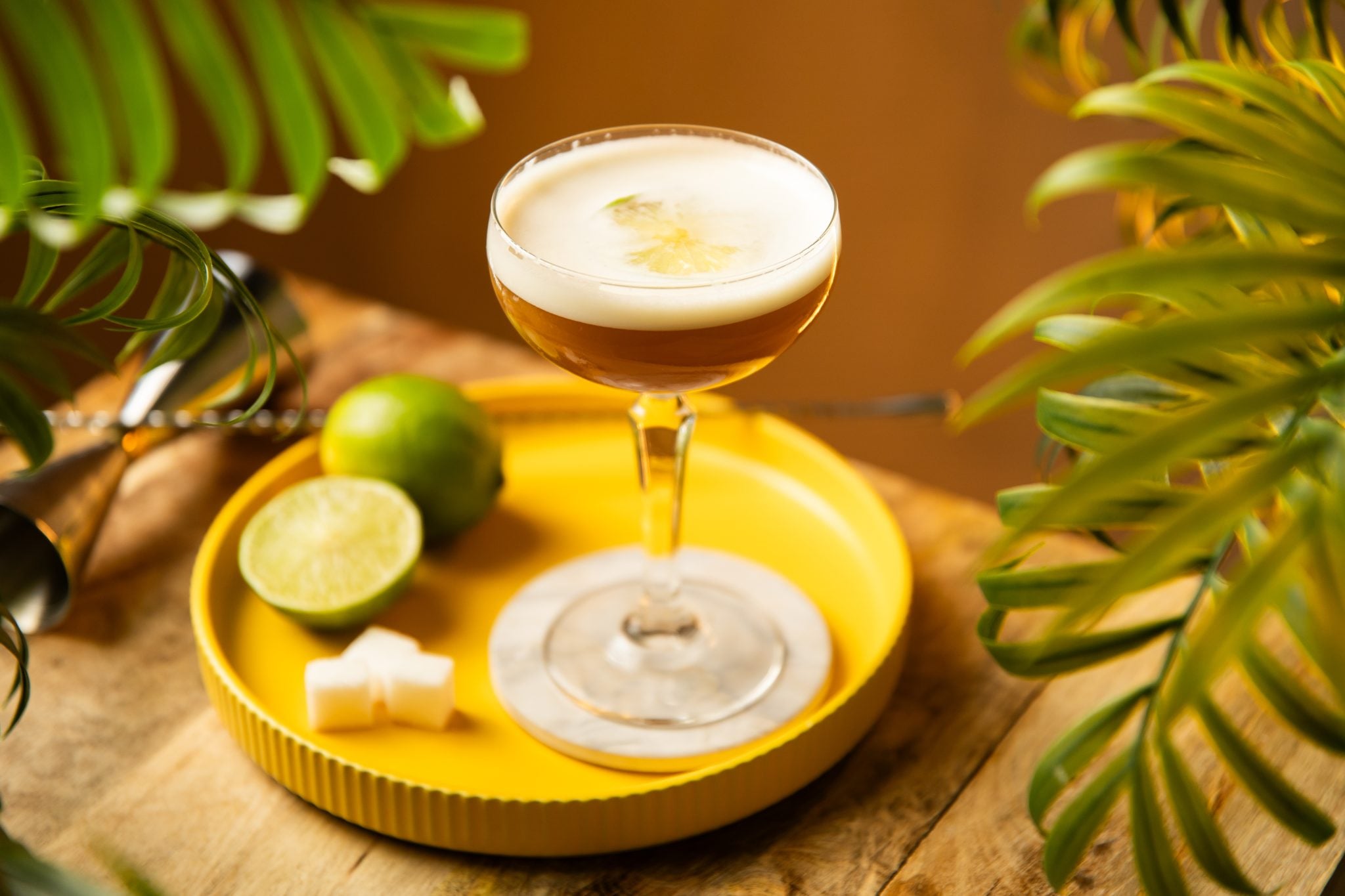 A side shot of a South of the Border cocktail in a coupe glass on a white coaster placed on a yellow tray surrounded by three sugar cubes, two limes, a bar spoon and a jigger