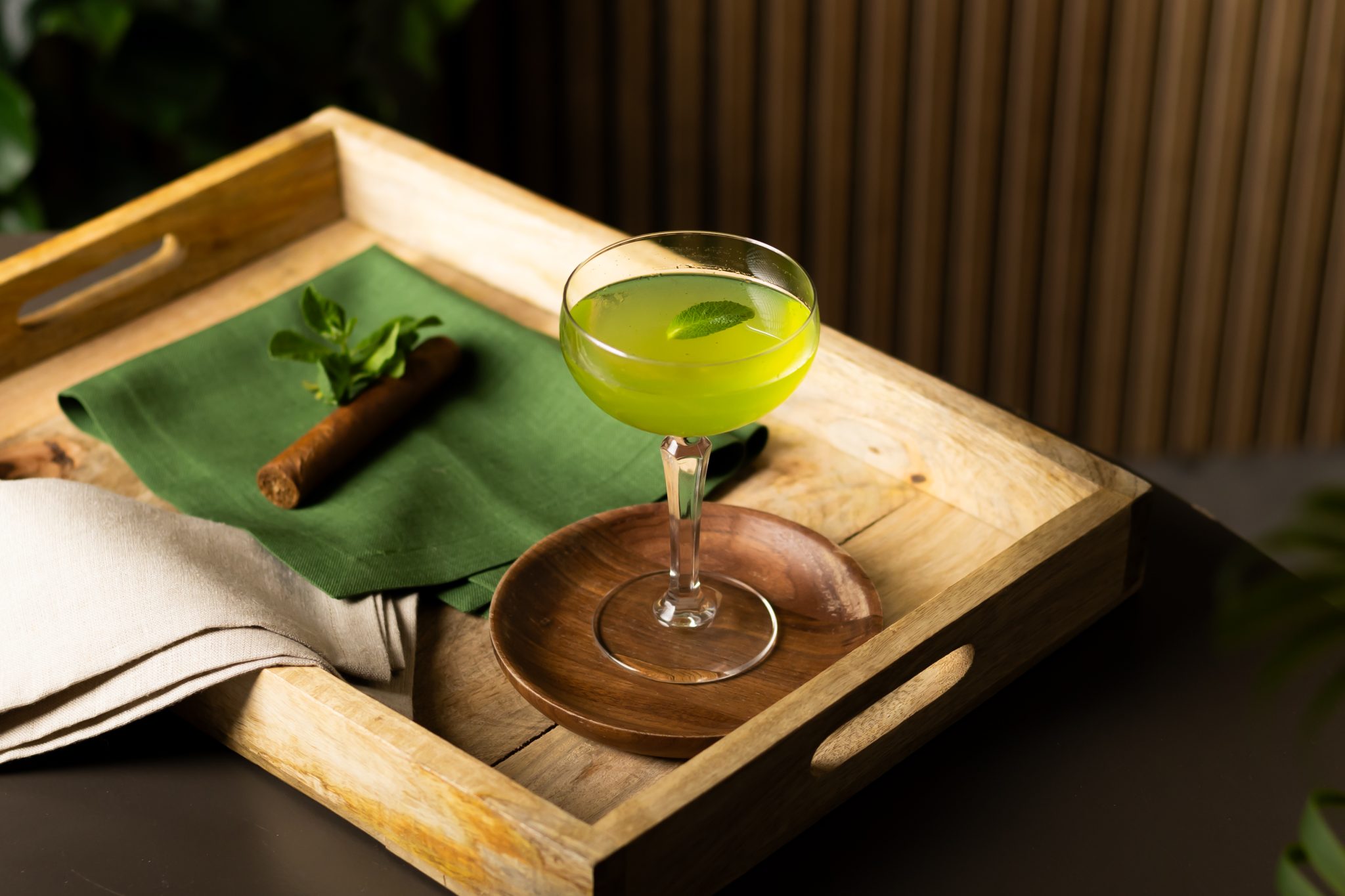 A side shot of a Southside cocktail in a martini glass on a wooden coaster placed on a wooden tray with a cigar, a green cloth and a beige cloth around.
