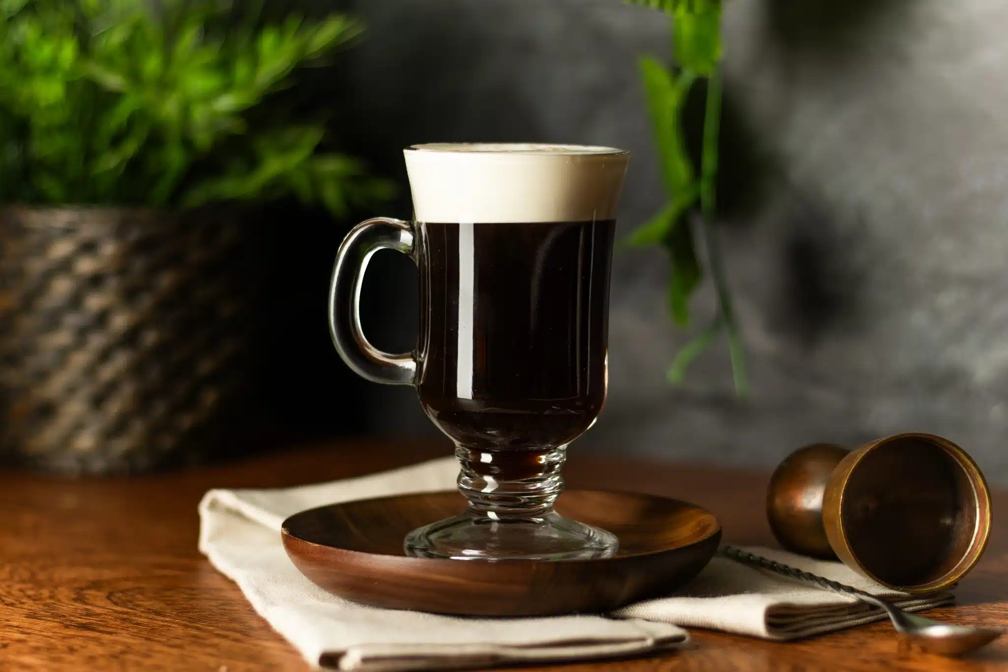 A side shot of a Spanish Coffee cocktail in a mug on a wooden plate placed on top of a beige cloth with a jigger and a bar spoon on the side, on top of a wooden table, in front of a plant and a dark grey stone wall.