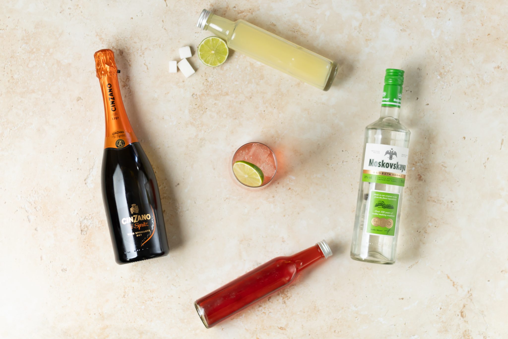 A Sparkling Cranberry Vodka Punch cocktail, shot from above, in a highball glasson a beige surface surrounded by Vodka, sparkling wine, cranberry juice, lime juice, simple syrup, surgar cubes and lime.