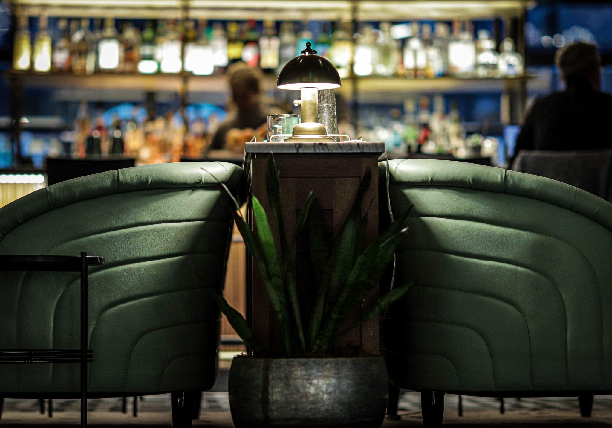 A shot of a Speakeasy bar showing two vintage chairs and a lamp with a bar table and spirits bottles behind