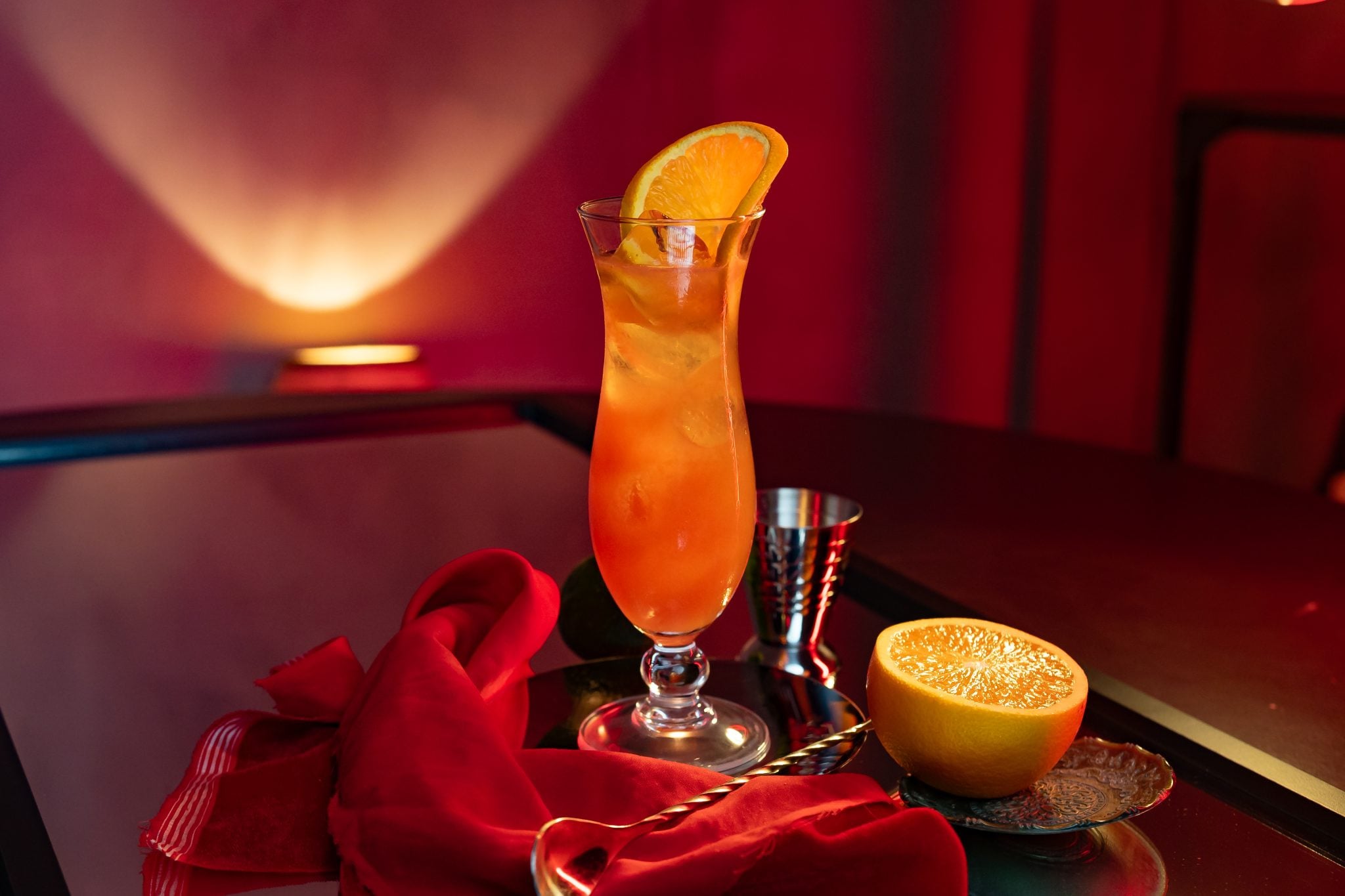 A side shot of a Spiced Hurricane cocktail in a hurricane glass on a wooden table surrounded by half a lemon, a jigger, a bar spoon, and a red cloth