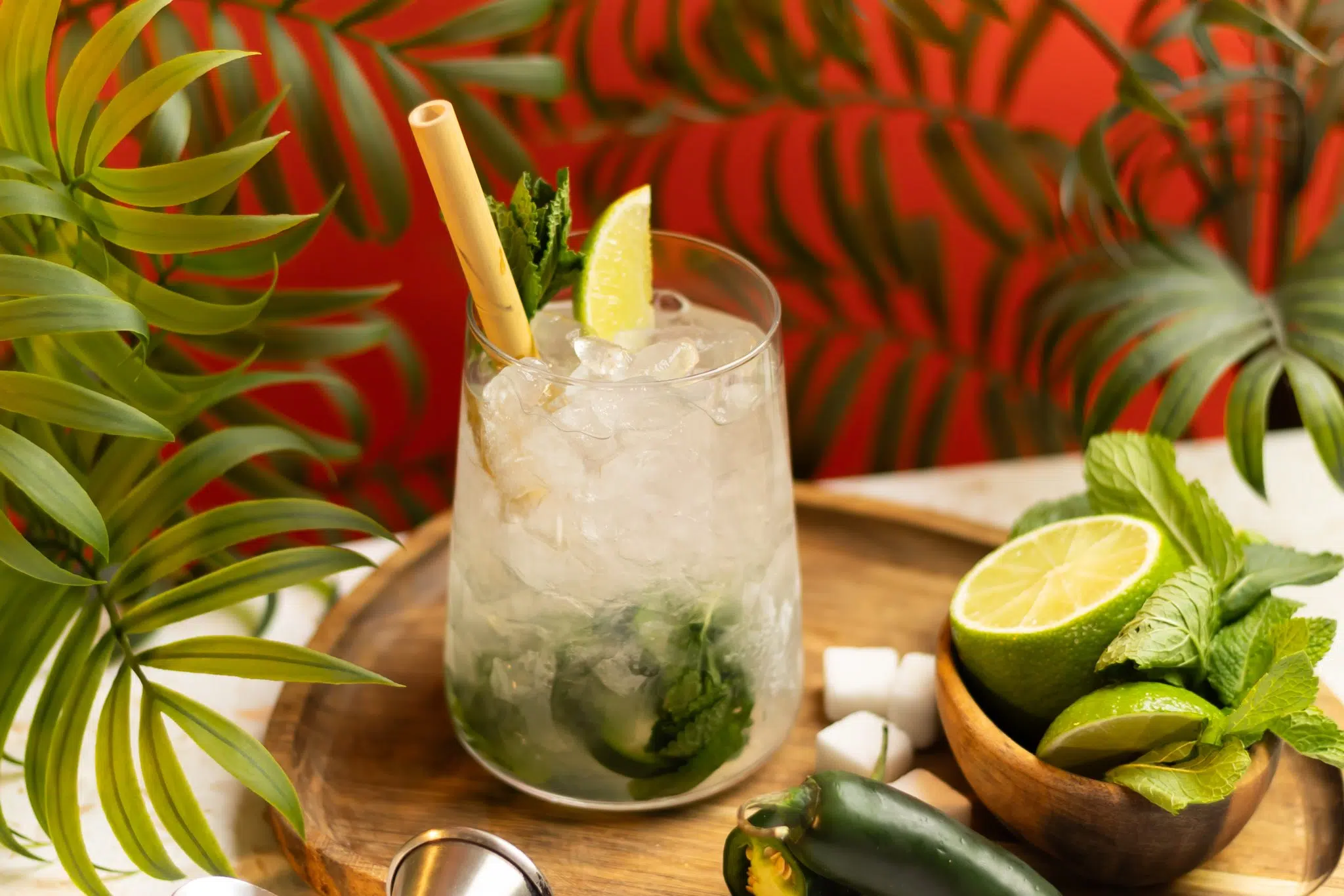 A side shot of a Spiced Mojito cocktail in highball glass on a wooden board surrounded by sugar cubes, a jalapeño and a bowl with limes and mint
