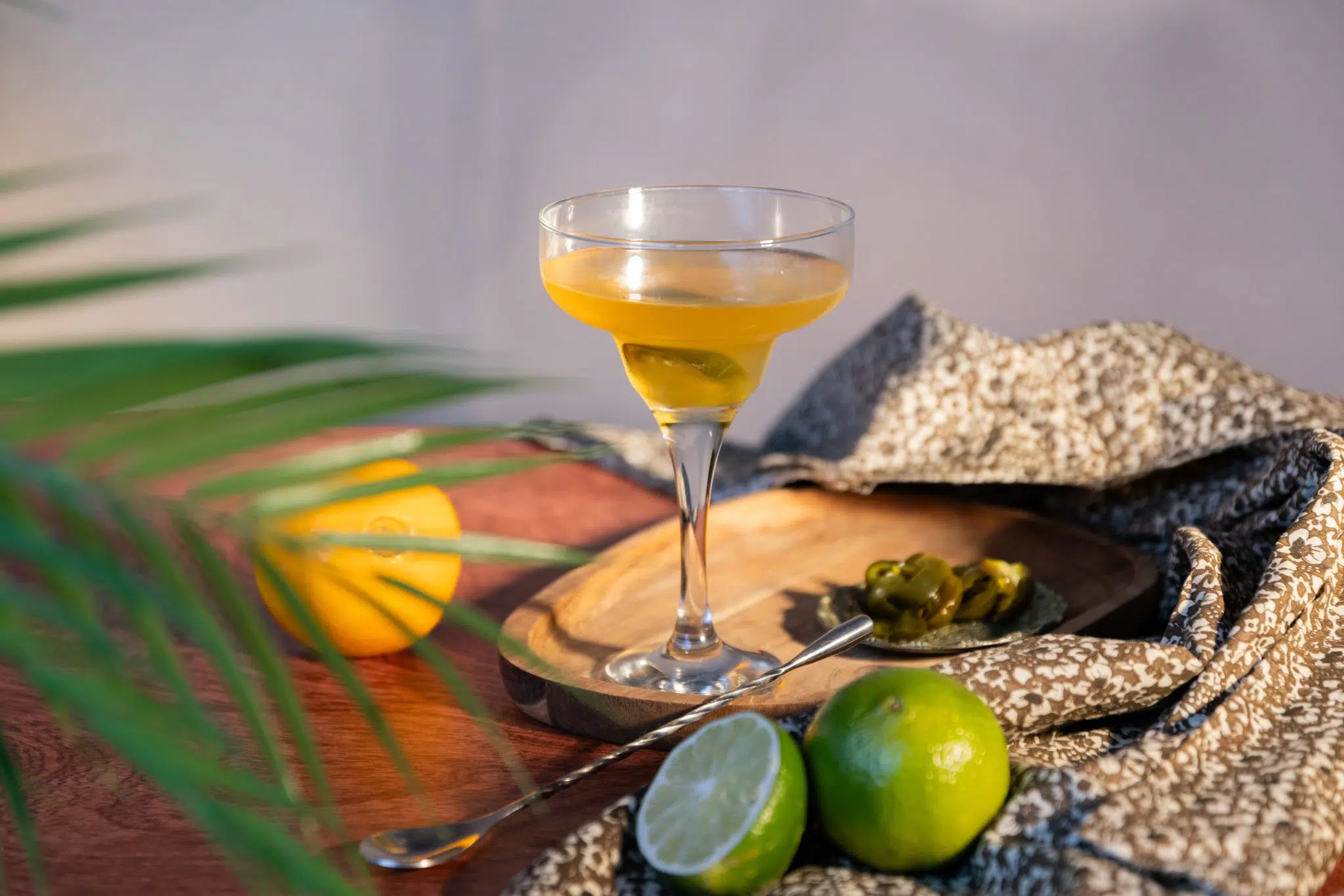 A side shot of a Spicy Margarita cocktail in a margarita glass on a wooden tray surrounded by two limes, a bar spoon, an orange, sliced jalapeño and a grey and white cloth