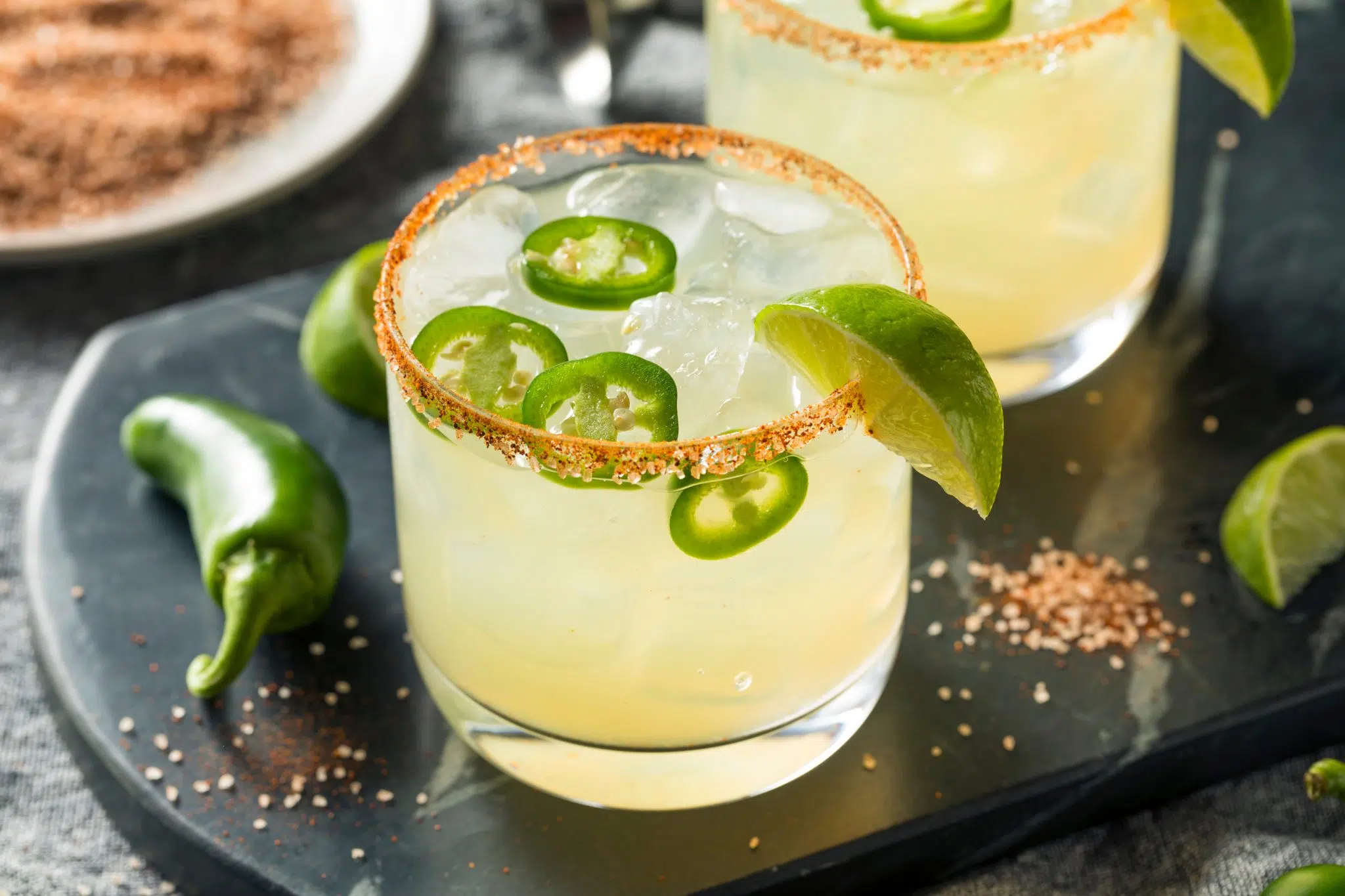 Two Spicy Margarita cocktails in low ball glasses on a black tray surrounded by a green chili pepper, a lime wedge, and red salt.