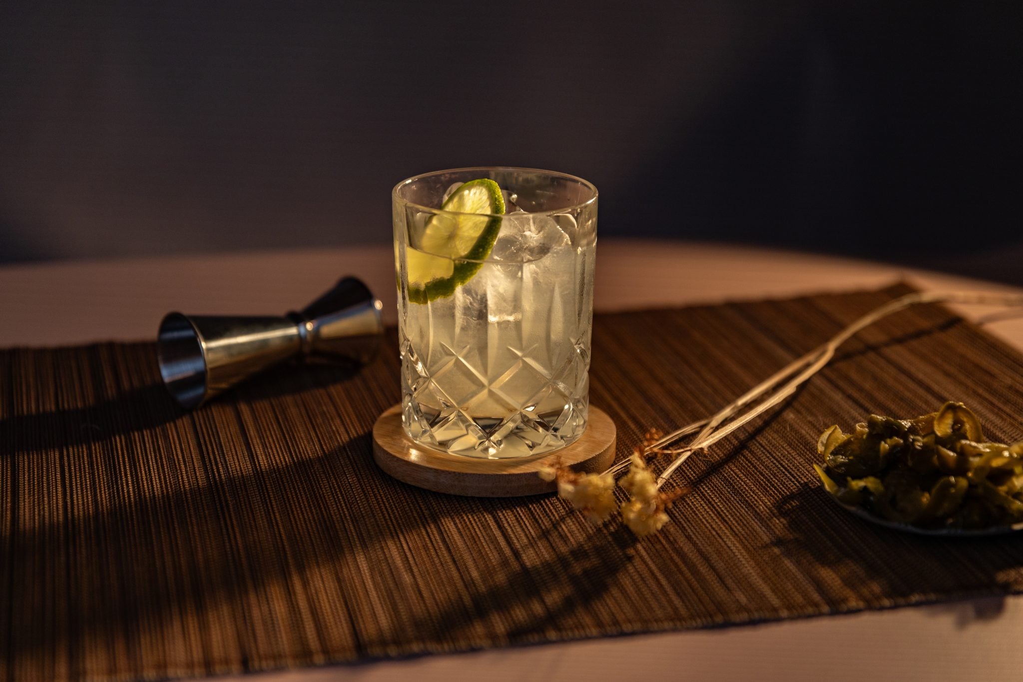 A side shot of a Spicy Soju Margarita cocktail in an old fashioned glass on a wooden coaster placed on a brown placemat surrounded by a jigger, a flower sprig and jalapeños slices