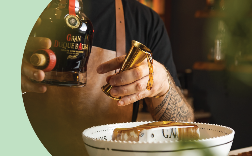 A shot of a bartender pouring a liquor with a jigger into a punch bowl on a huge ice cube