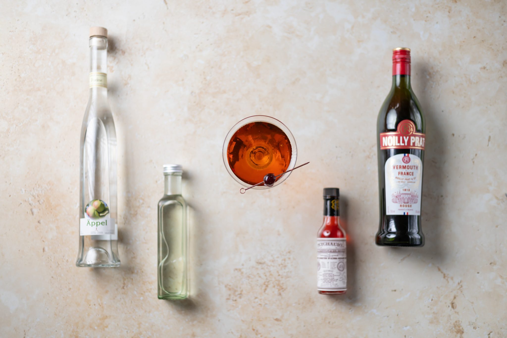 A Star cocktail, shot from above, in a martini glass on a beige surface surrounded by apple brandy, sweet vermouth, gomme syrup, and Peychaud's bitters