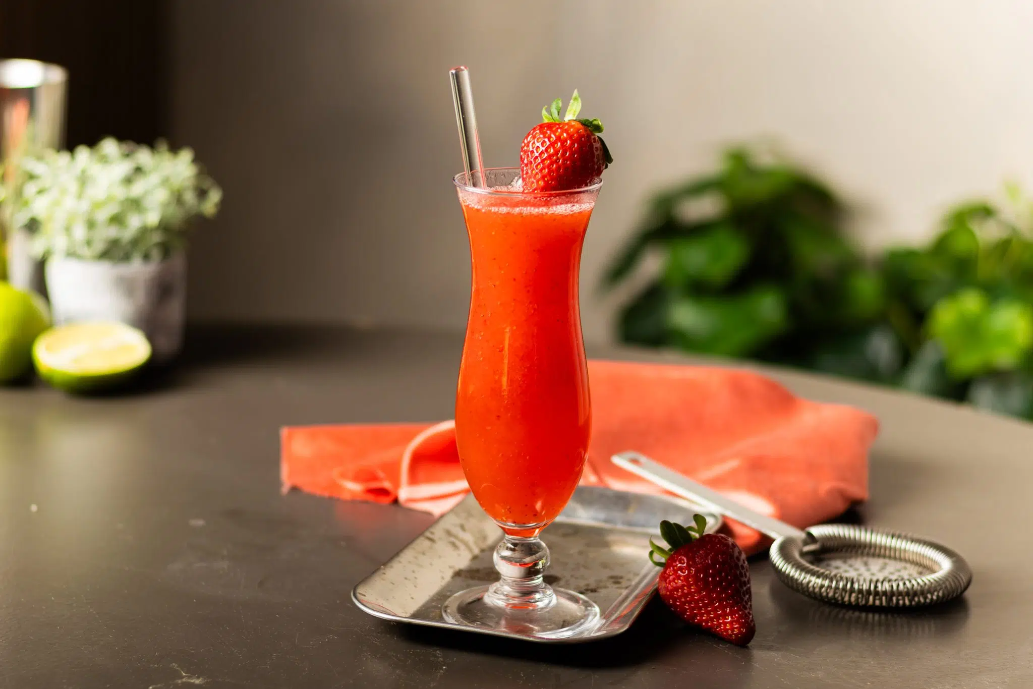 A side shot of a Strawberry Margarita in a hurricane glass on a silver metal tray on a black table surrounded by a strainer, a red cloth and a strawberry.