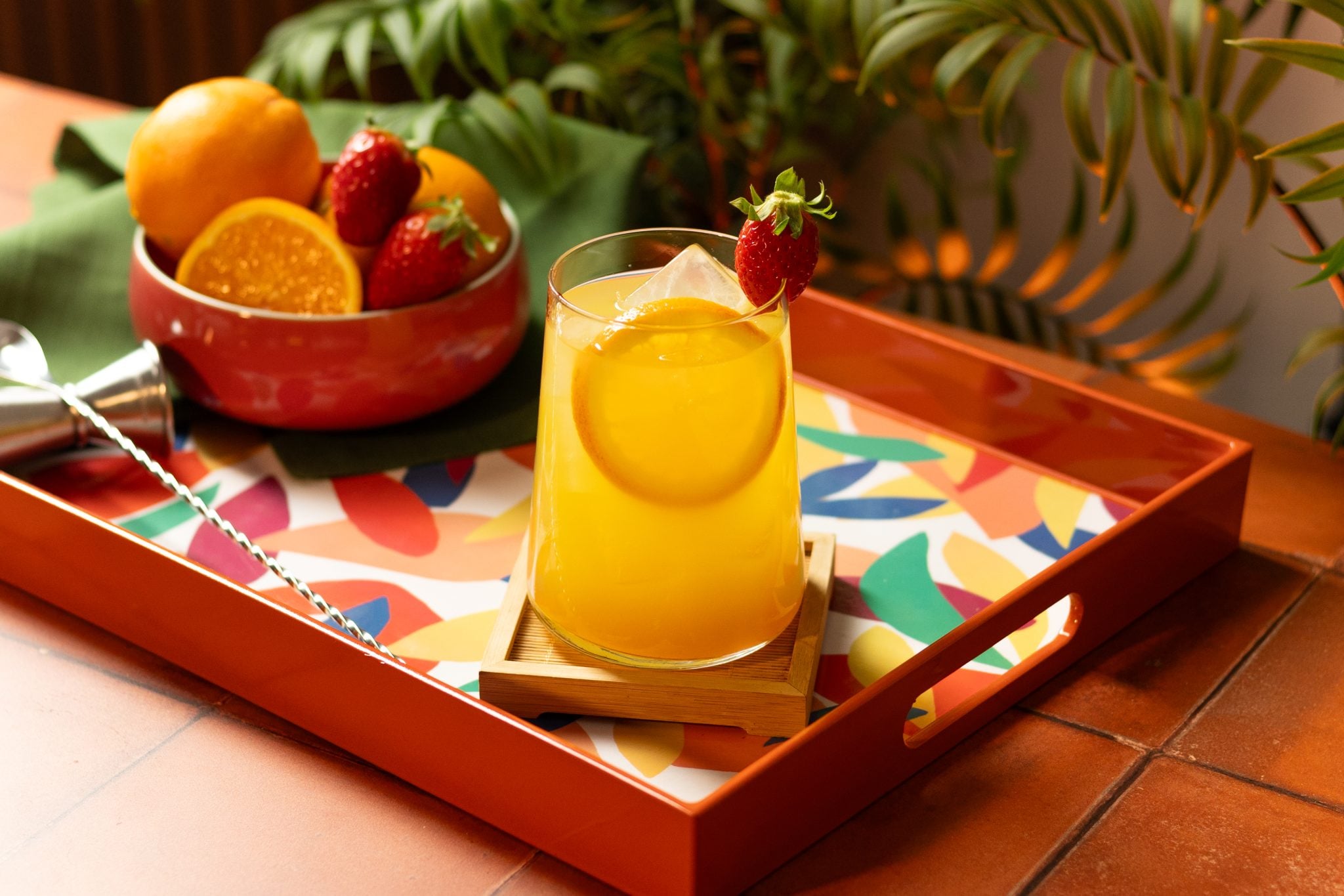 A side shot of a Strawberry Tequila Sunrise cocktail in a highball glass on a wooden coaster placed on a multicolor tray with a oranges and strawberries bowl , a bar spoon and a green cloth around