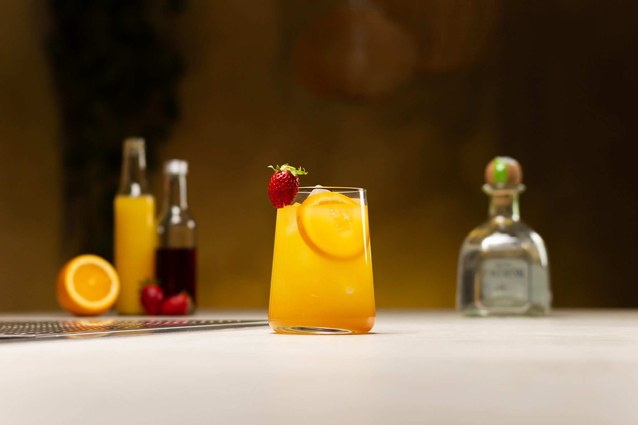 Tequila, orange juice, orange, strawberry syrup, and strawberries laid out on a white bar table