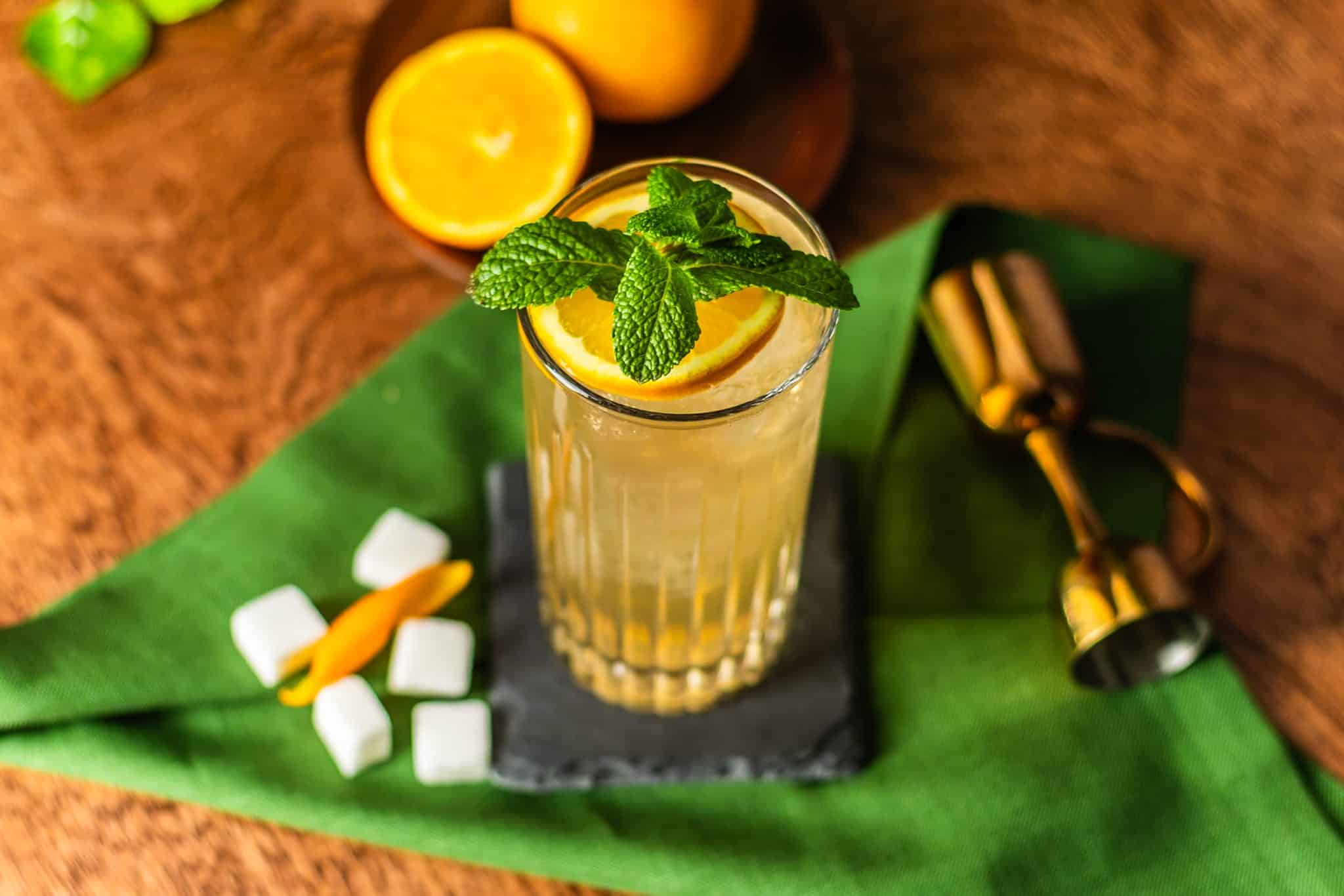 A Suffering Bastard cocktail, shot from above, in highball glass on black stone coaster on a green cloth placed on a wooden table with sugar cubes, orange pieces, and a jigger around.